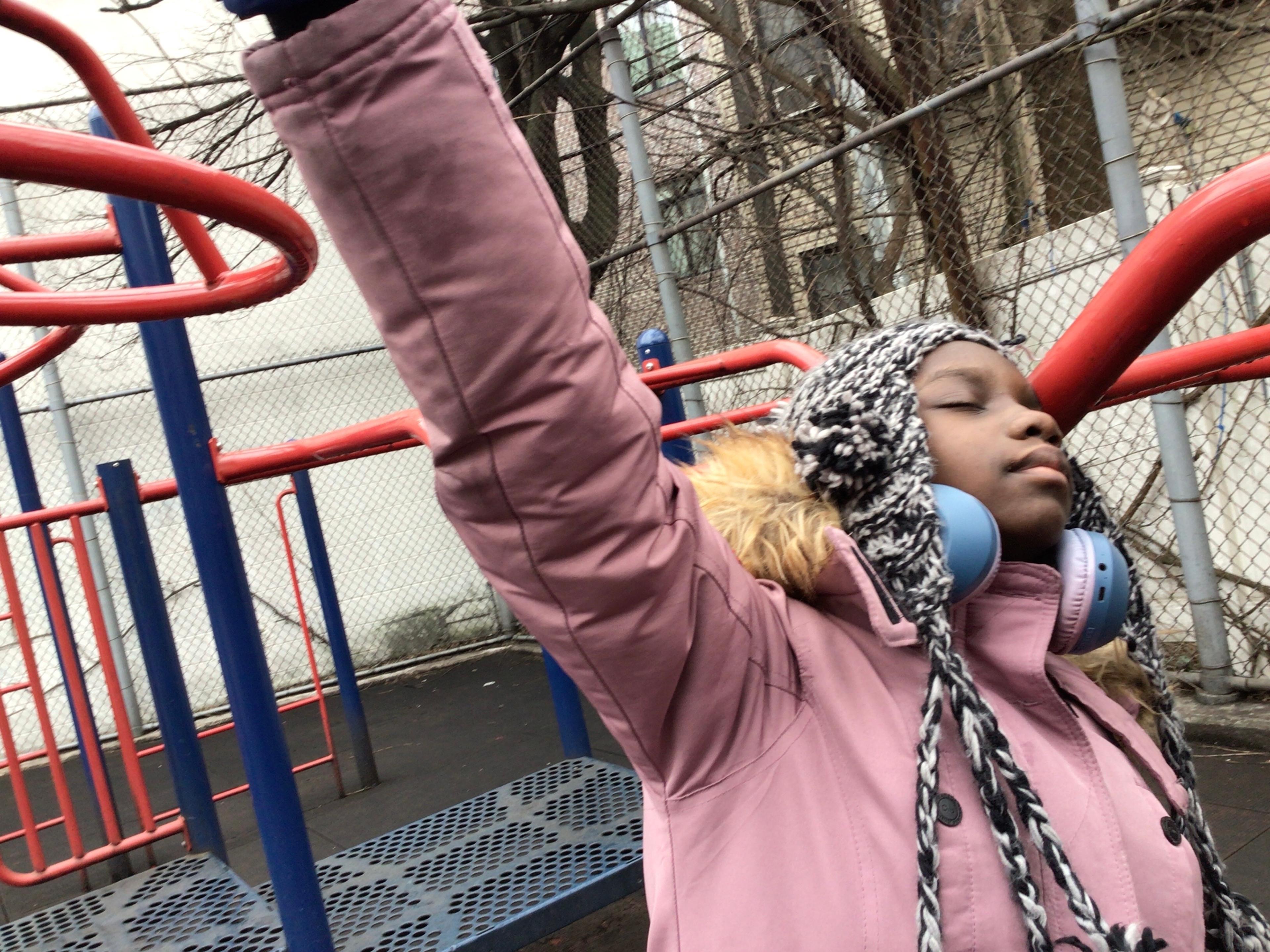 Color digital photograph of a young Black girl standing in the foreground of an outdoor playground on an overcast day. She faces right and wears a heavy pink coat. Her right arm is raised up and extends out of the frame at top. Her eyes are closed and her head is tilted upward and back. She wears big blue headphones around her neck, and a black and white braided wool cap with earflaps and tassels on her head. Behind the girl is a large playground set made of blue and red iron bars, with tall chainlink fences to the rear at the corners of the playground.