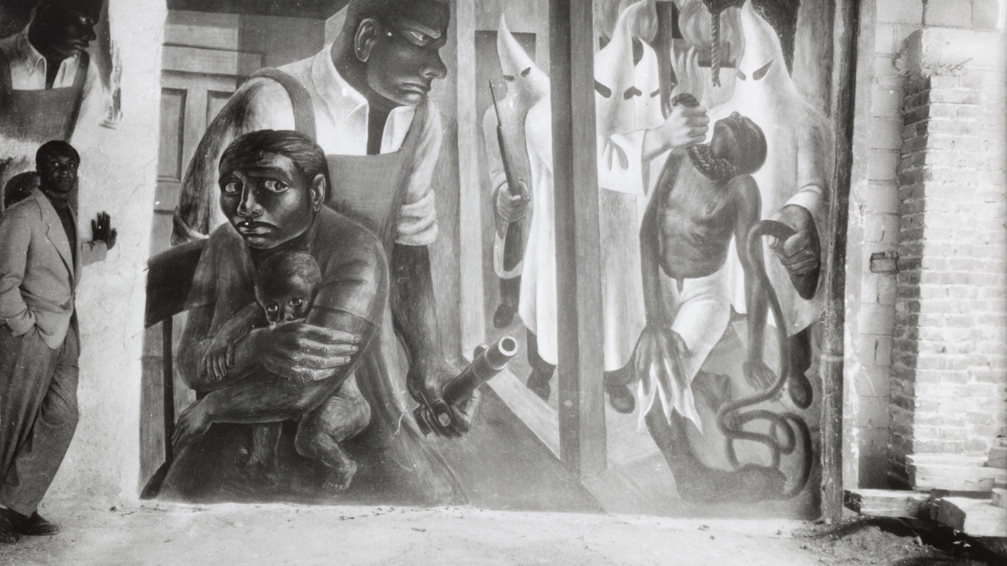 Black and white image of man standing next to mural depicted in black and white of a mother shielding her baby and while a strong black meale guards her. 