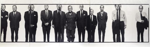 Eleven stoic men stand side-by-side. The man in the center wears military fatigues, while the rest wear suits.