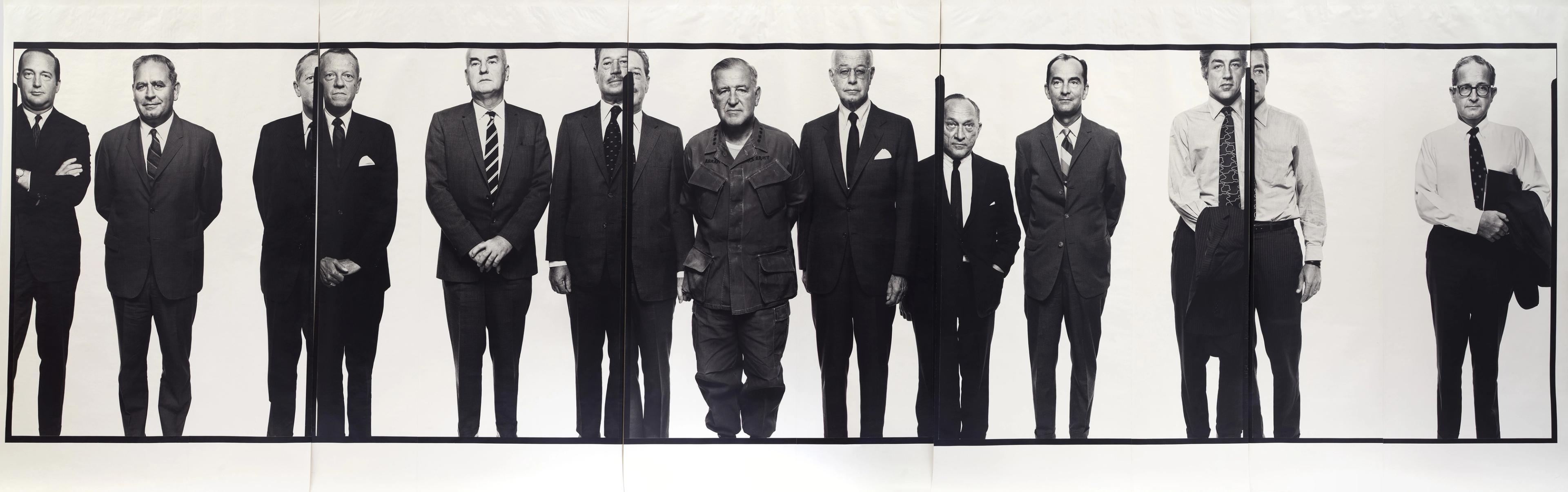 Eleven stoic men stand side-by-side. The man in the center wears military fatigues, while the rest wear suits.