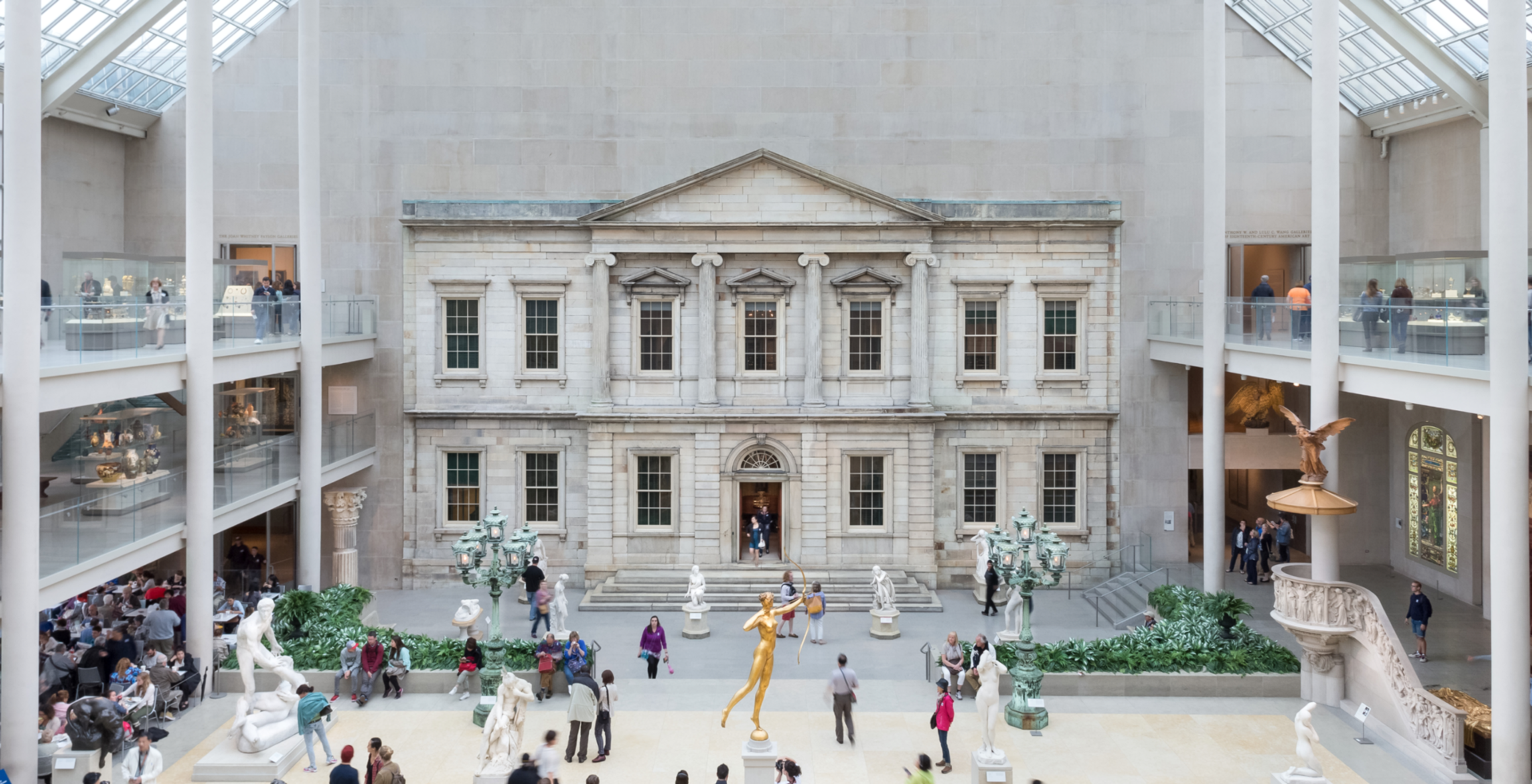 Photo of the American Wing gallery at The Metropolitan museum of art: an indoor courtyard with sky lights with the facade of a bank vault on one wall with people roaming around the courtyard.