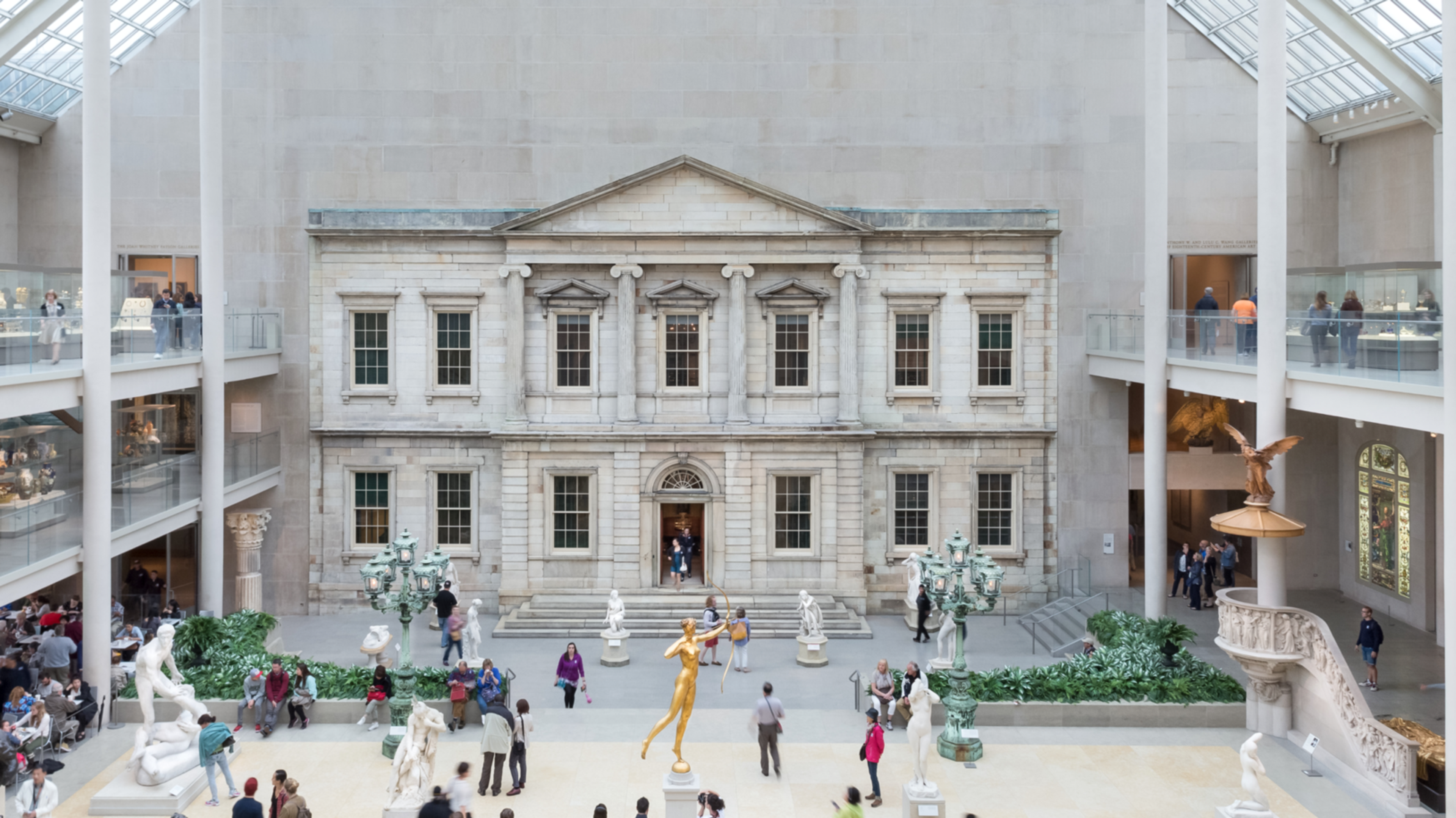 Photo of the American Wing gallery at The Metropolitan museum of art: an indoor courtyard with sky lights with the facade of a bank vault on one wall with people roaming around the courtyard.