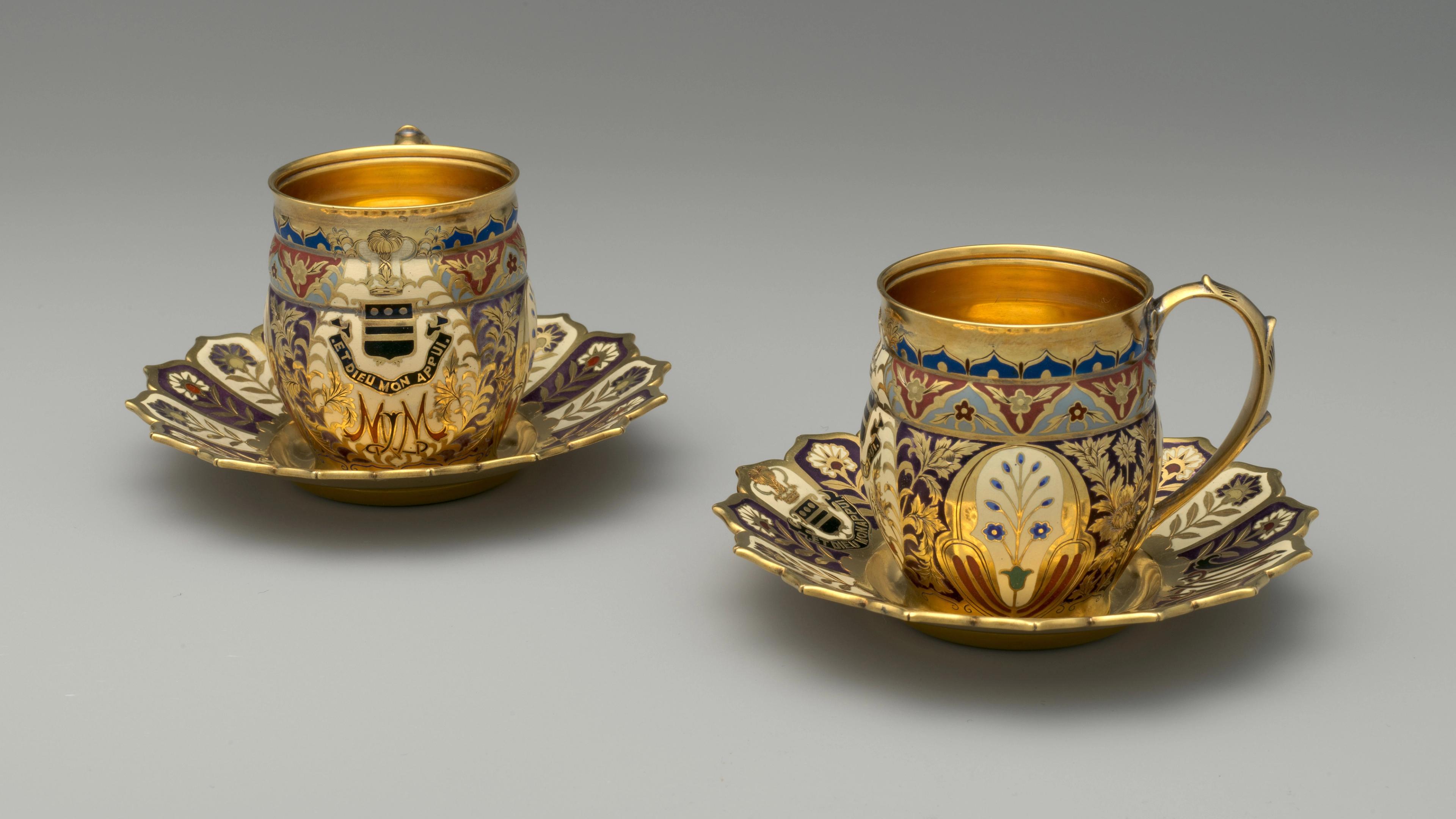 A pair of dazzling gilded and enameled cups and saucers adorned with floral designs, a coat of arms, and monogram.