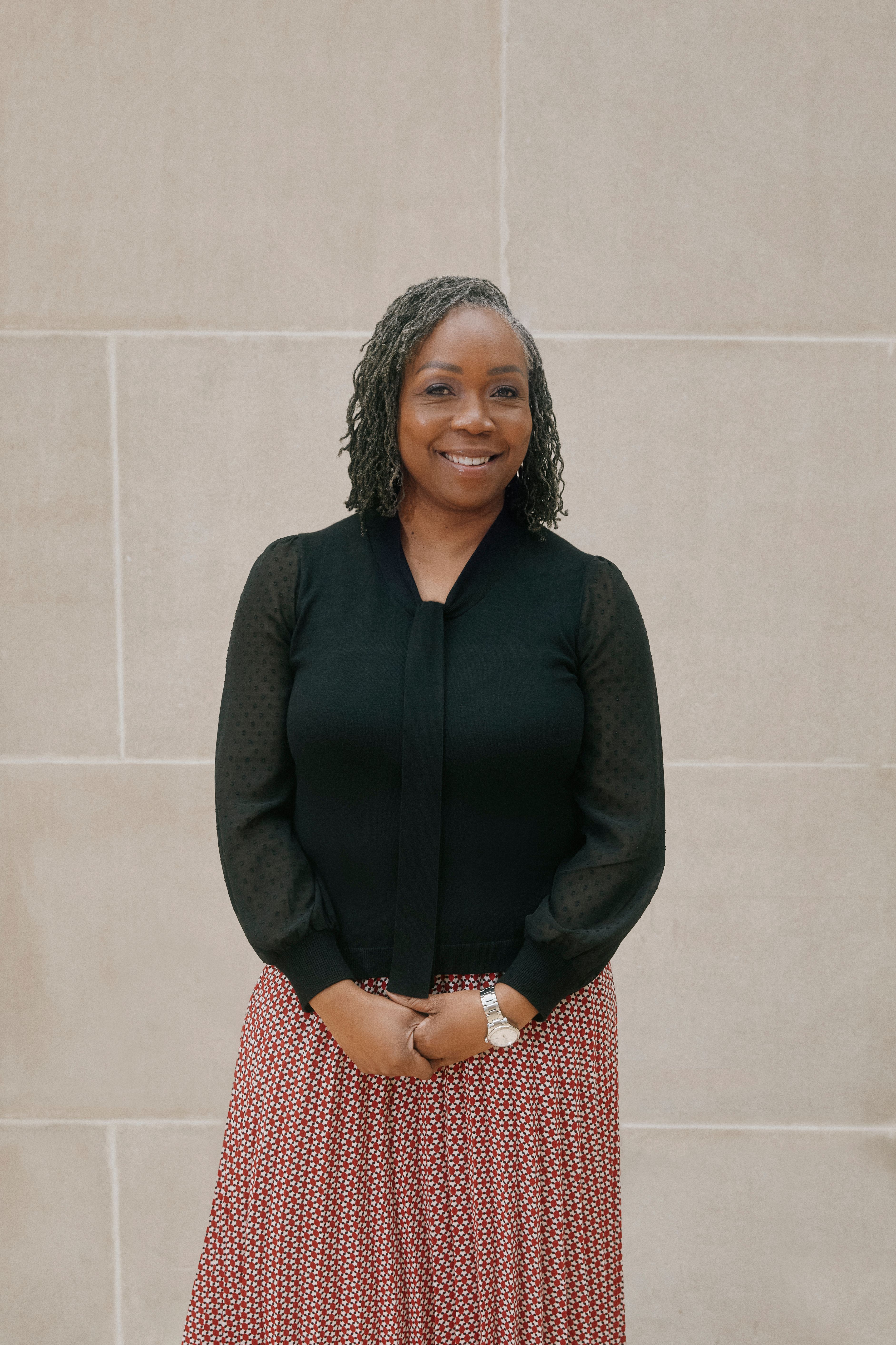 A color photograph of Lavita McMath Turner standing in front of a stone wall in a black top and red and white patterned skirt.