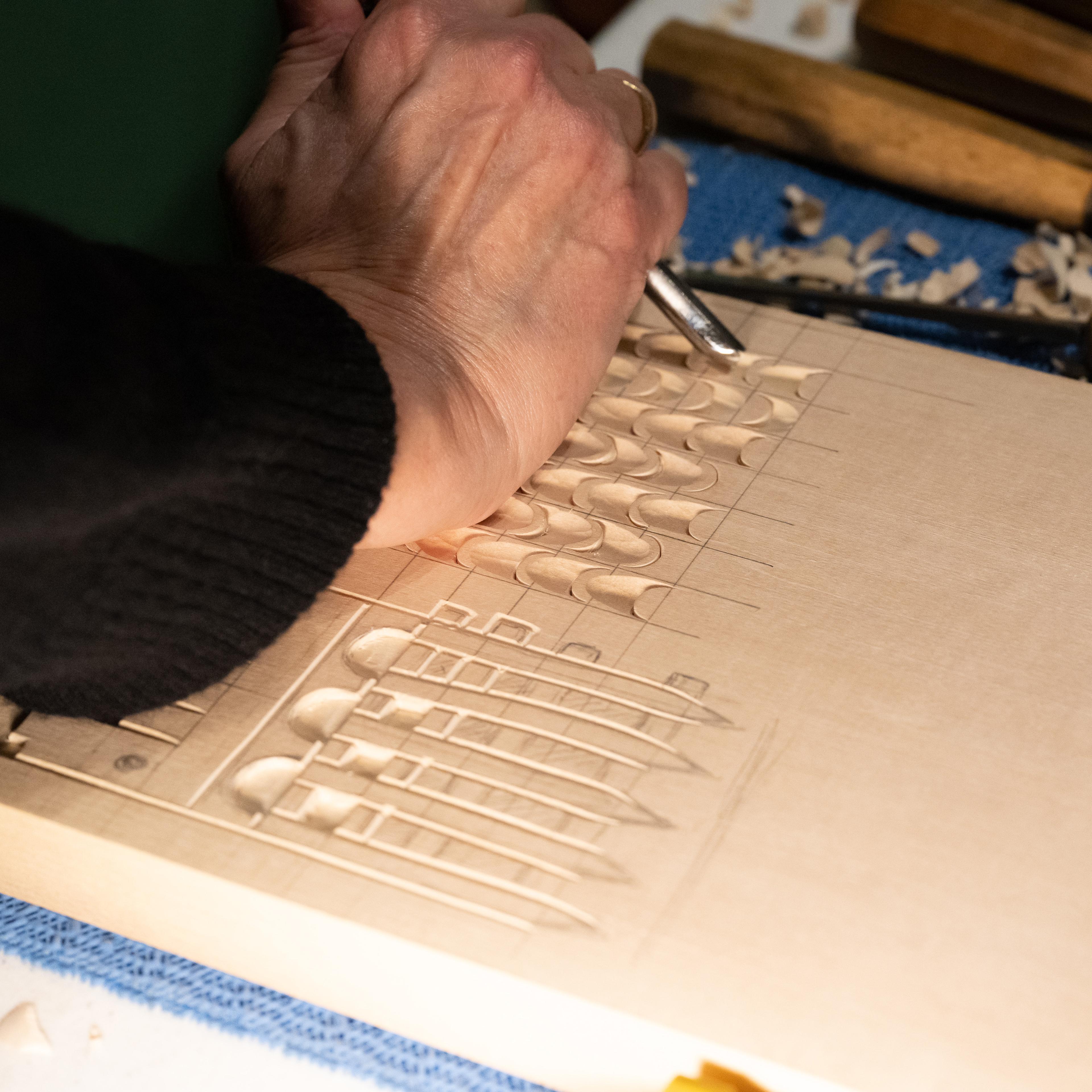 A hand holds a wood carving tool. The tool is being used to carve a design that looks like waves on a wooden board.