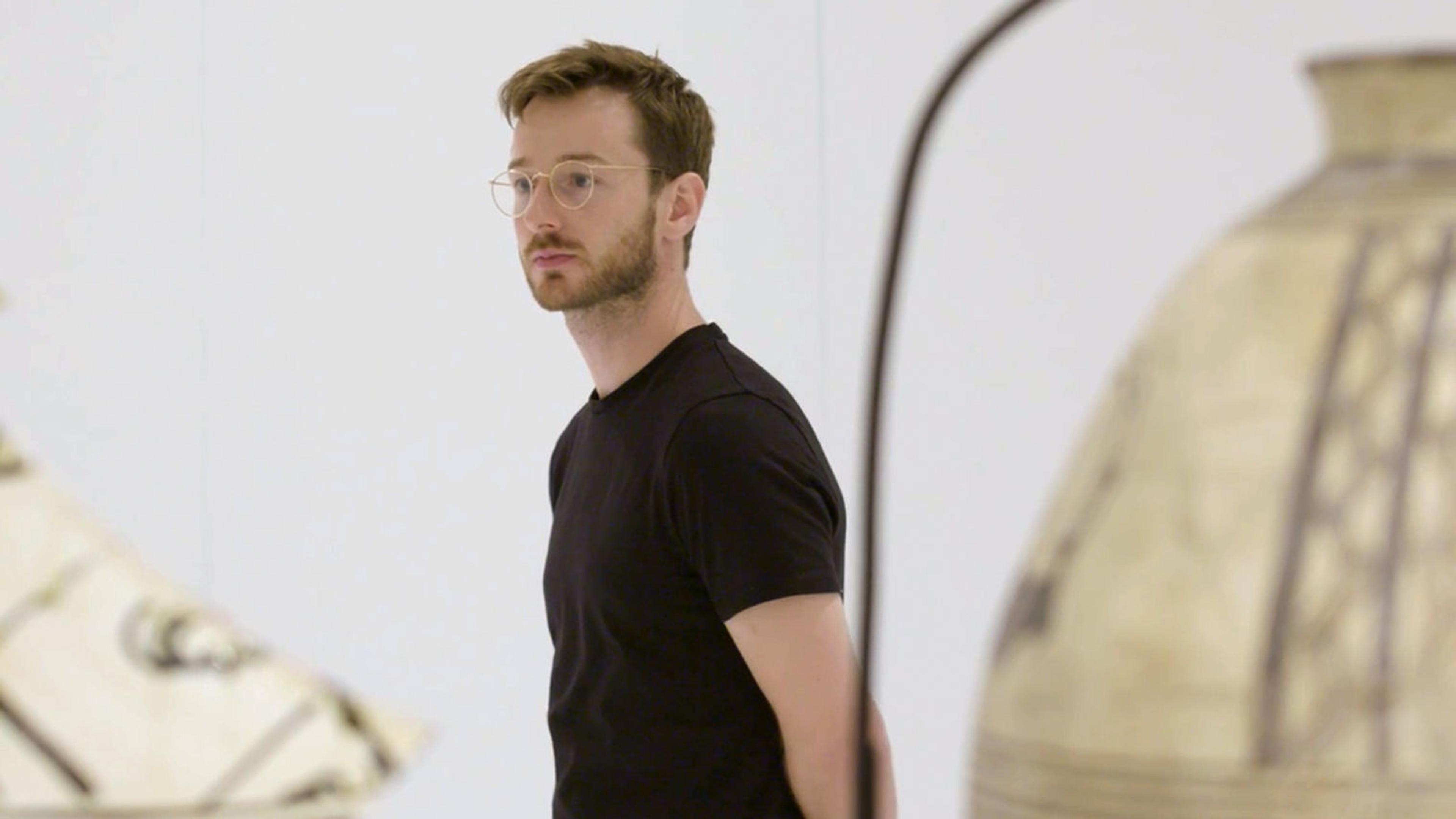 A blonde-haired man with glasses, dressed in black, stands between two sculptures