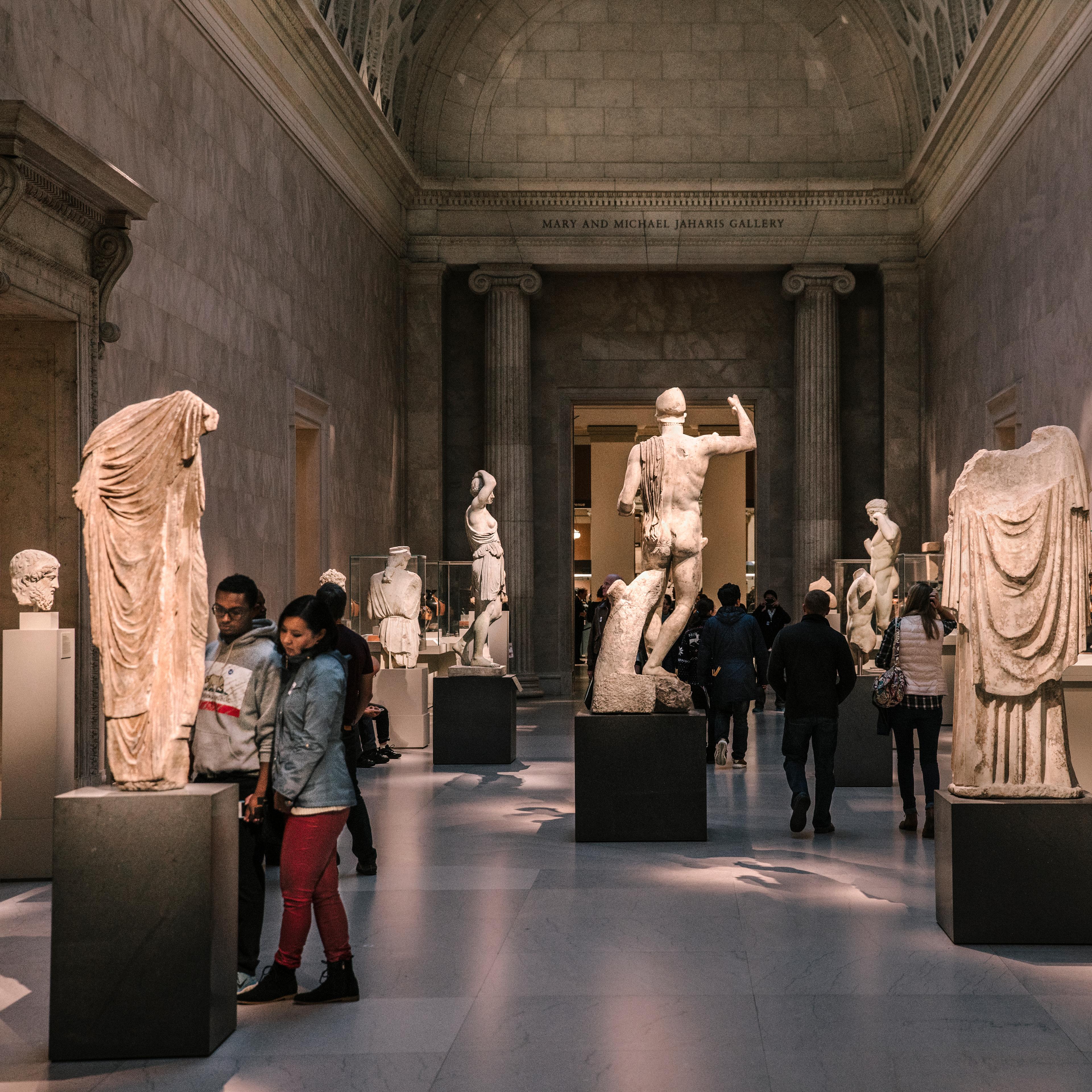 Visitors are in a Greek and Roman gallery at The Met that is filled with large white statues of figures. It's evening and the room is dark with spotlights on the art.  