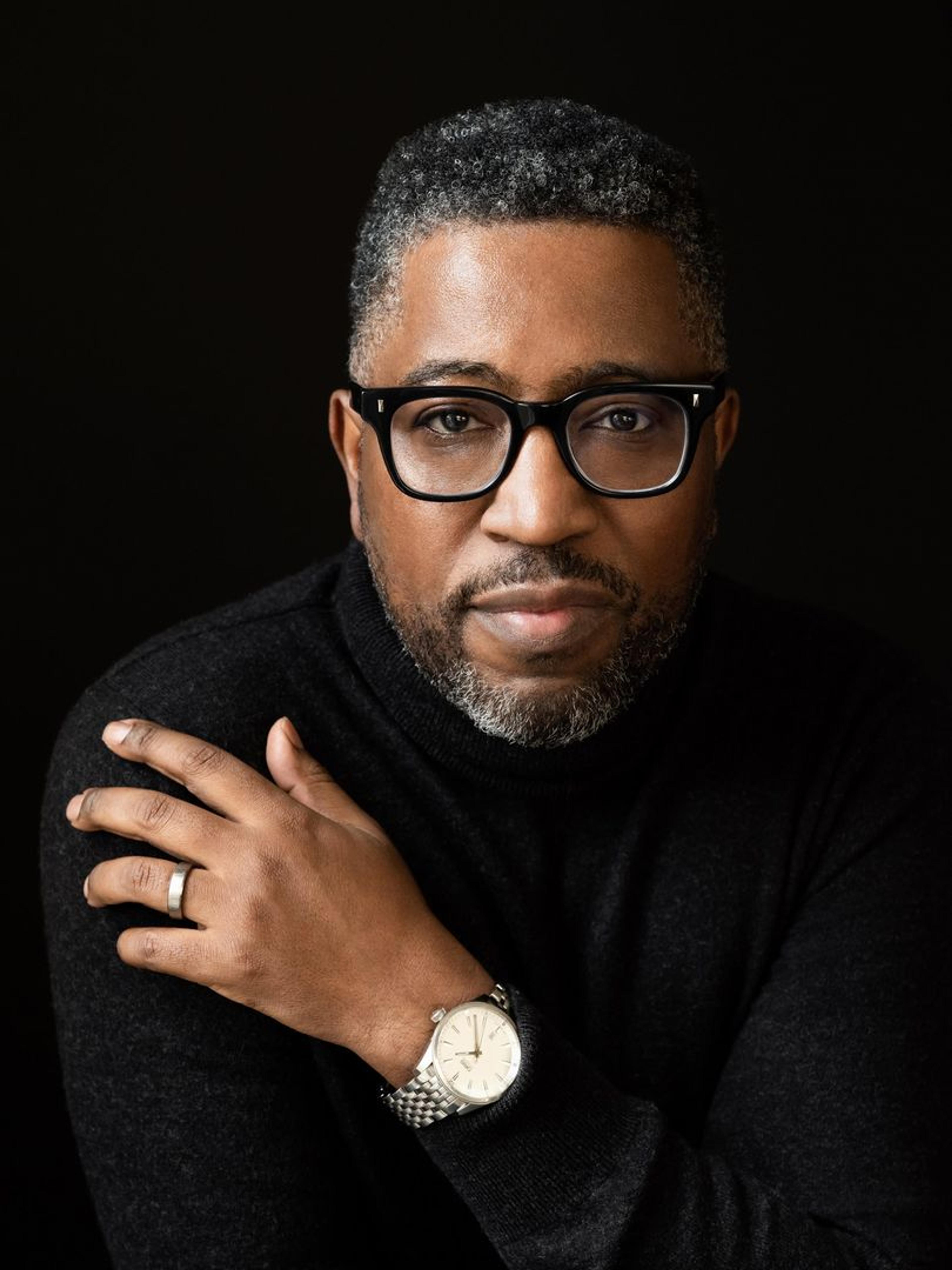 A portrait of a man with glasses wearing a black turtleneck and a watch, posing with his hand on his shoulder against a black background.