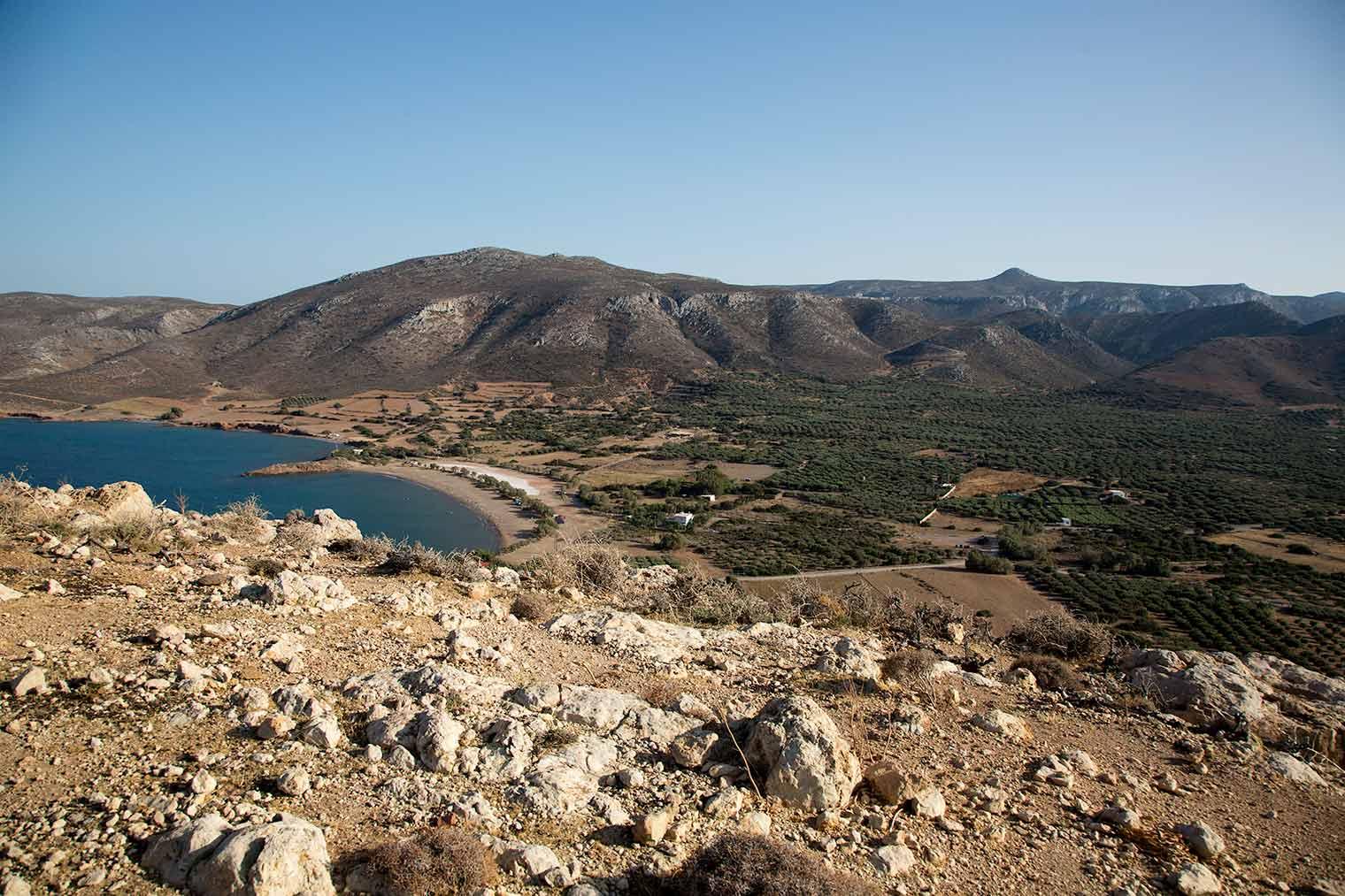 A scenic coastal landscape view from a rocky hillside, overlooking a curved bay and farmland with rugged mountains in the background under a clear blue sky.