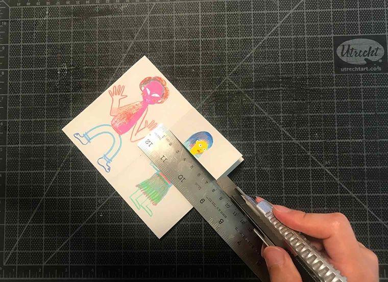 The folded paper is laid flat on the cutting board. The ruler sits over the top third of the paper while I hand slices the paper with a craft knife.