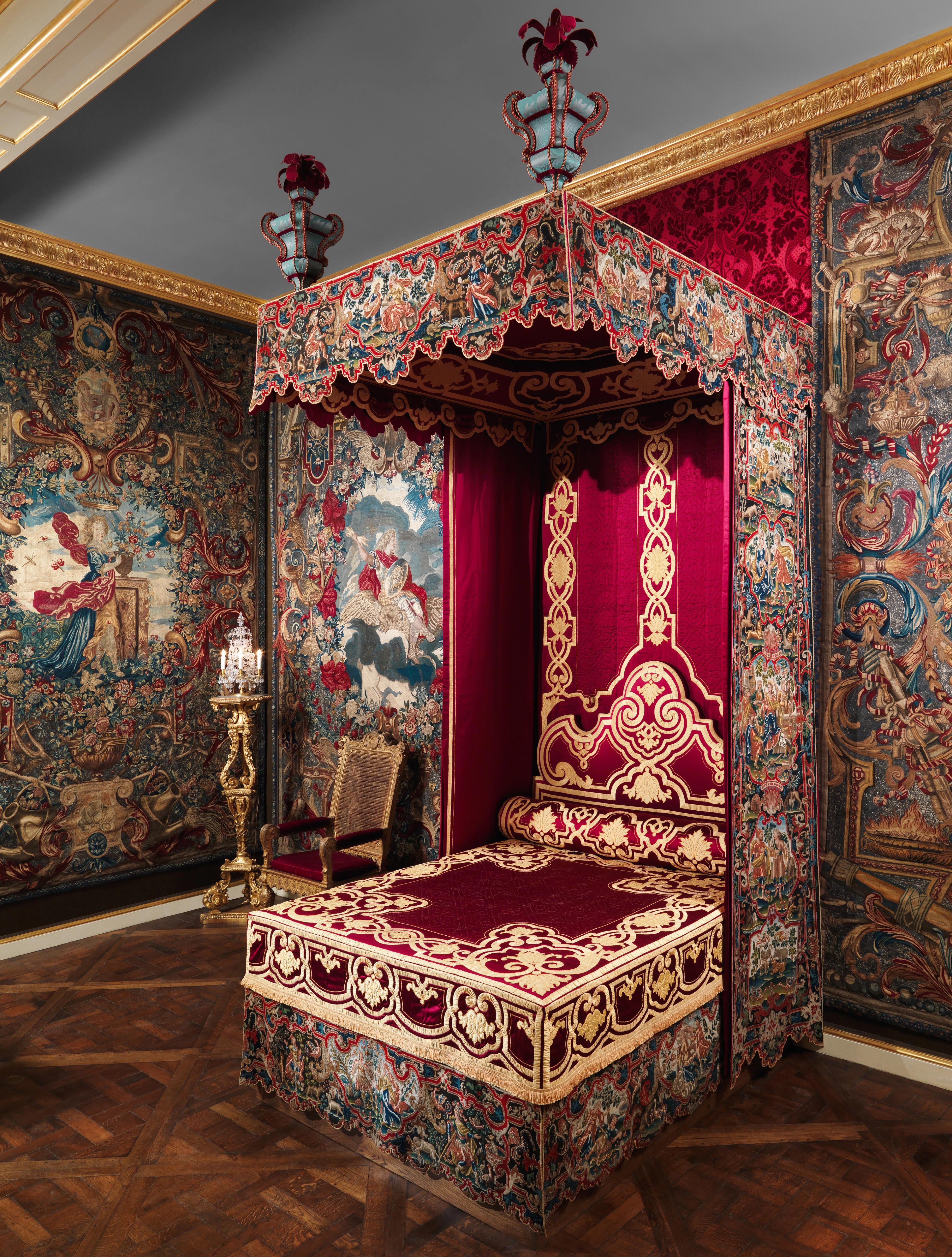 Ornate canopied bed upholstered in dark red velvet with gold trim, displayed in a gallery surrounded by large decorative tapestries hanging on the walls.