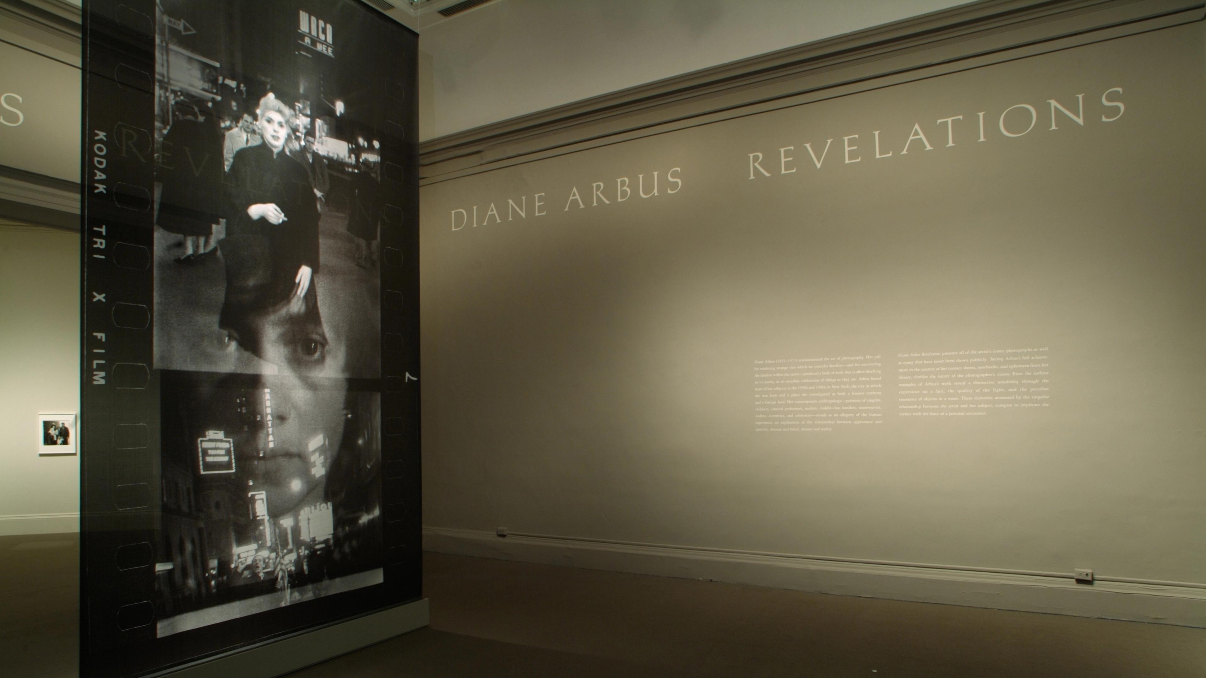 Dimly lit gallery with a stanchion displaying photo contact sheets in the foreground, and large text reading "DIANE ARBUS REVELATIONS" along the far wall.
