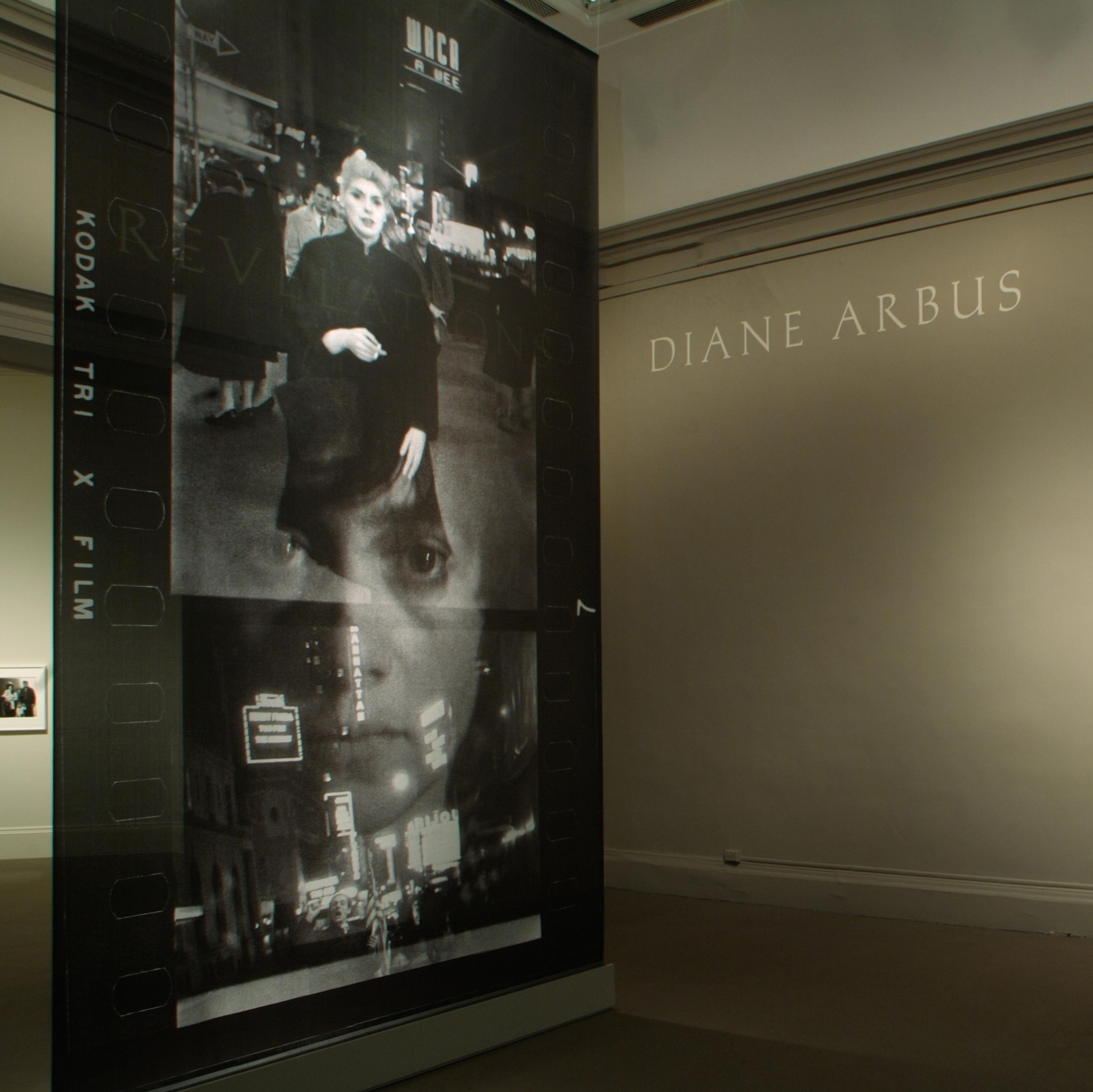 Dimly lit gallery with a stanchion displaying photo contact sheets in the foreground, and large text reading "DIANE ARBUS REVELATIONS" along the far wall.