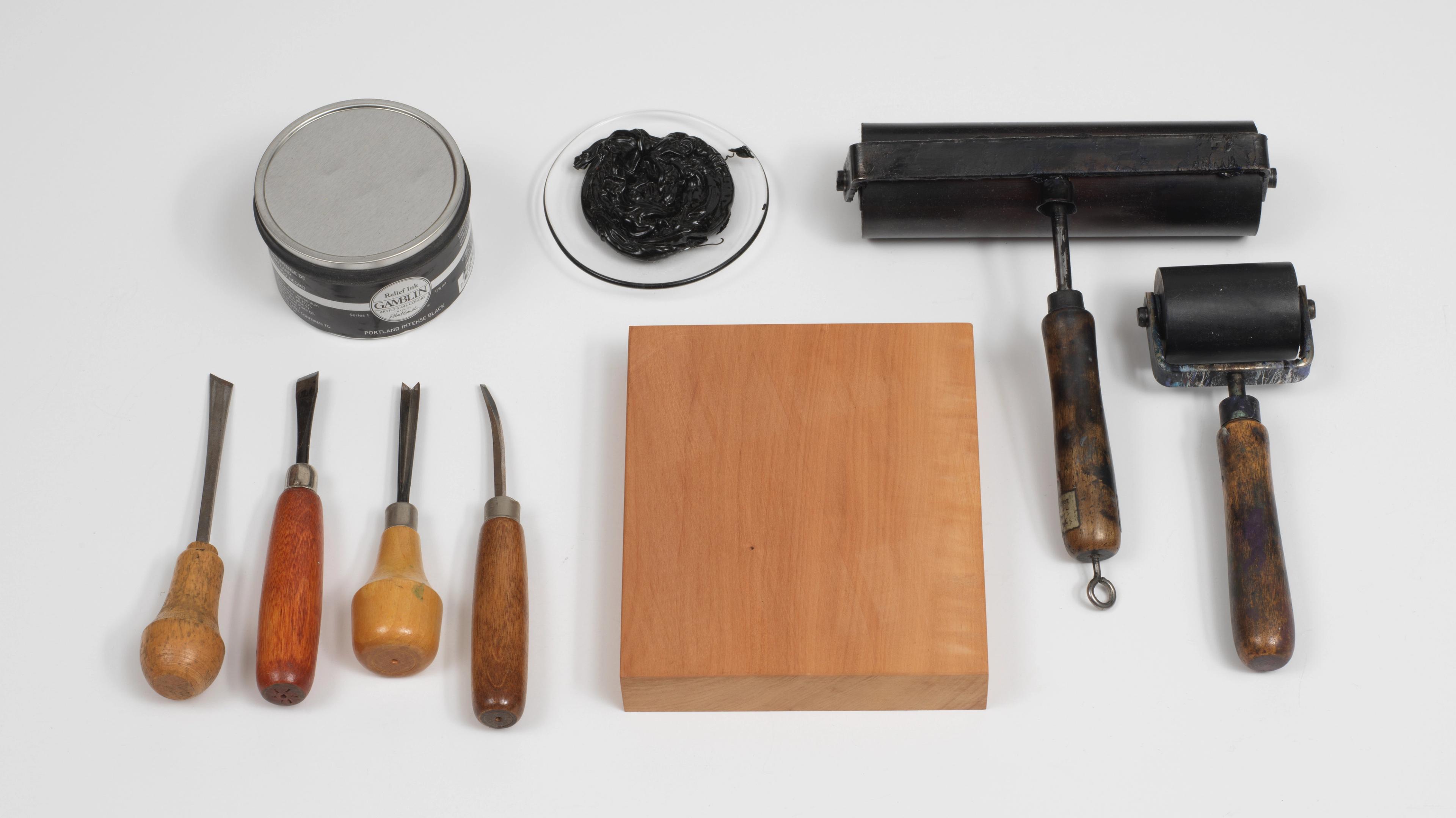 Photo of woodcutting tools, including a block, chisel, gouche, knife, ink ball, roller, and ink.
