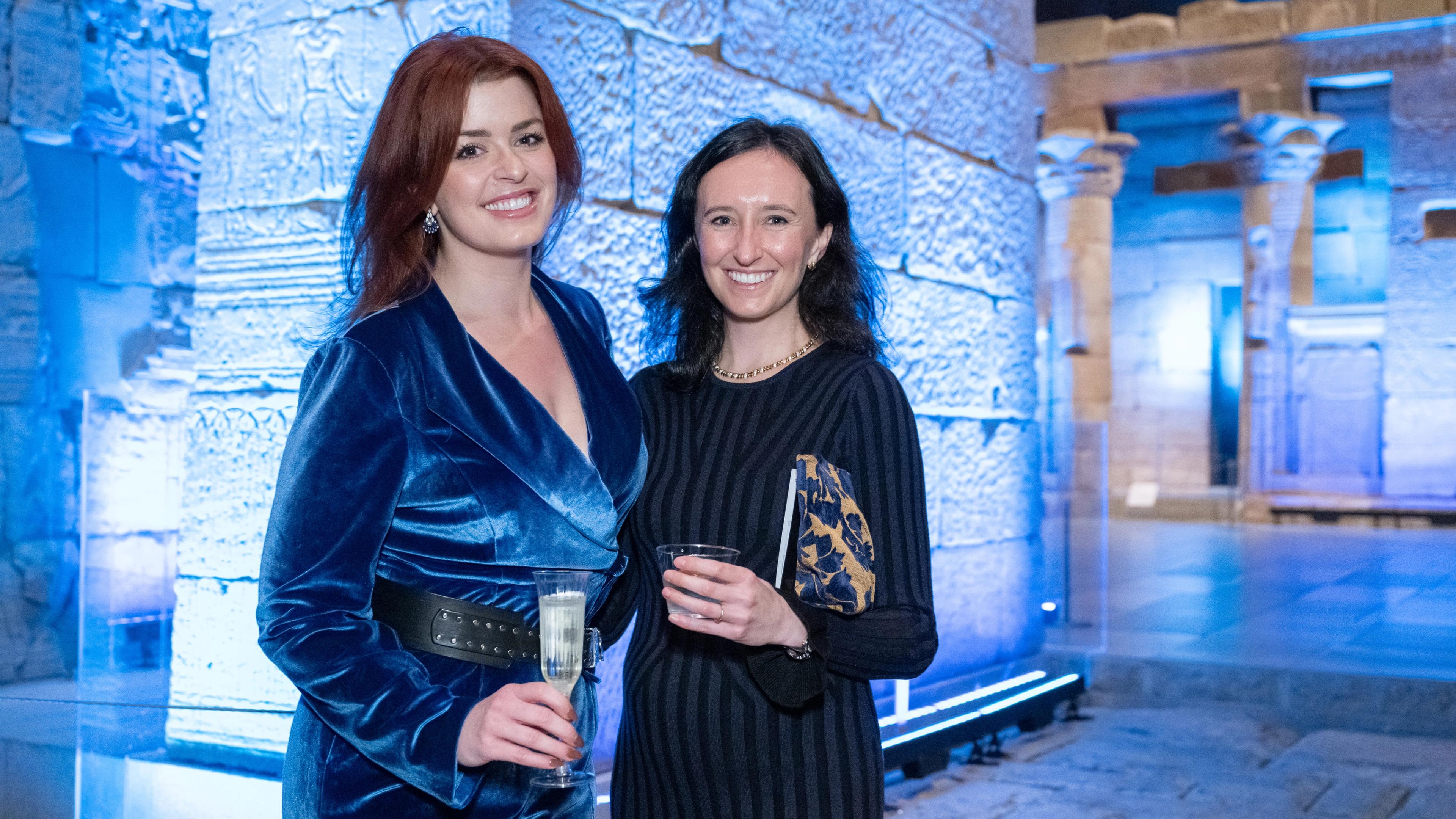 Two women stand in front of the Temple of Dendur at The Metropolitan Museum of Art, holding cocktails and smiling.
