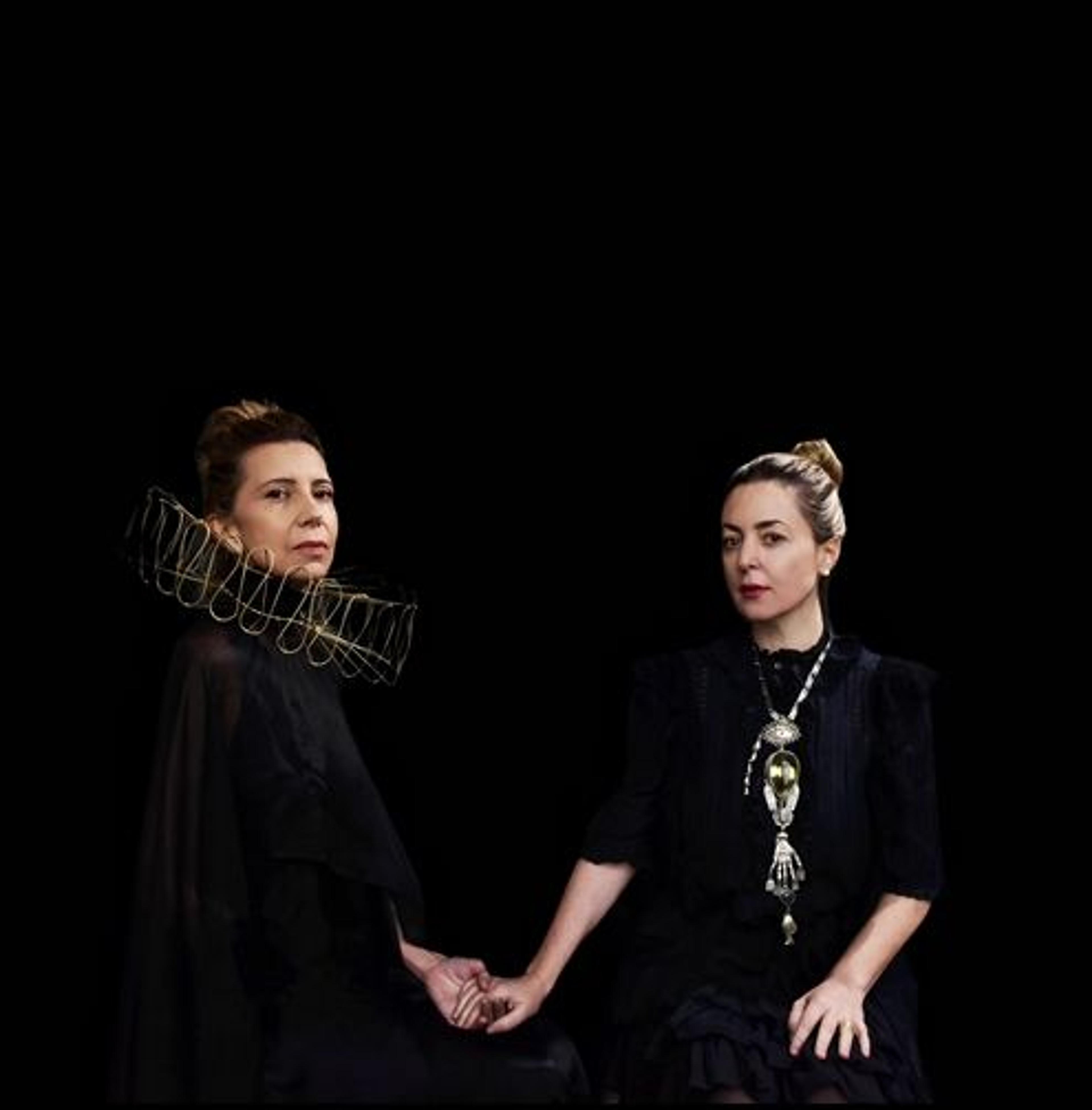 Two women against a solid backdrop loosely holding hands. The one on the left has a ruffled collar and the one on the right has a decorative necklace