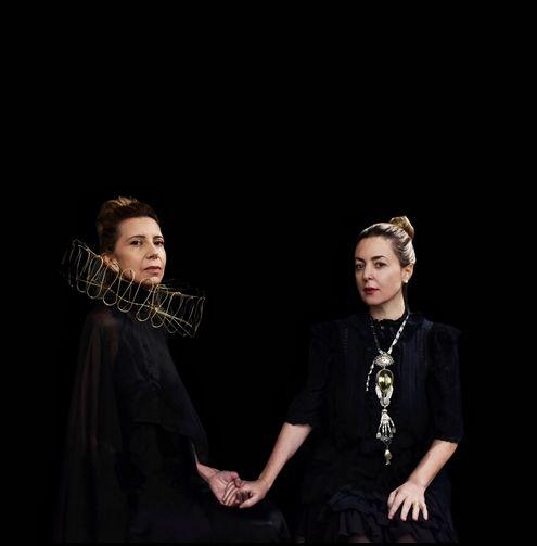 Two women against a solid backdrop loosely holding hands. The one on the left has a ruffled collar and the one on the right has a decorative necklace