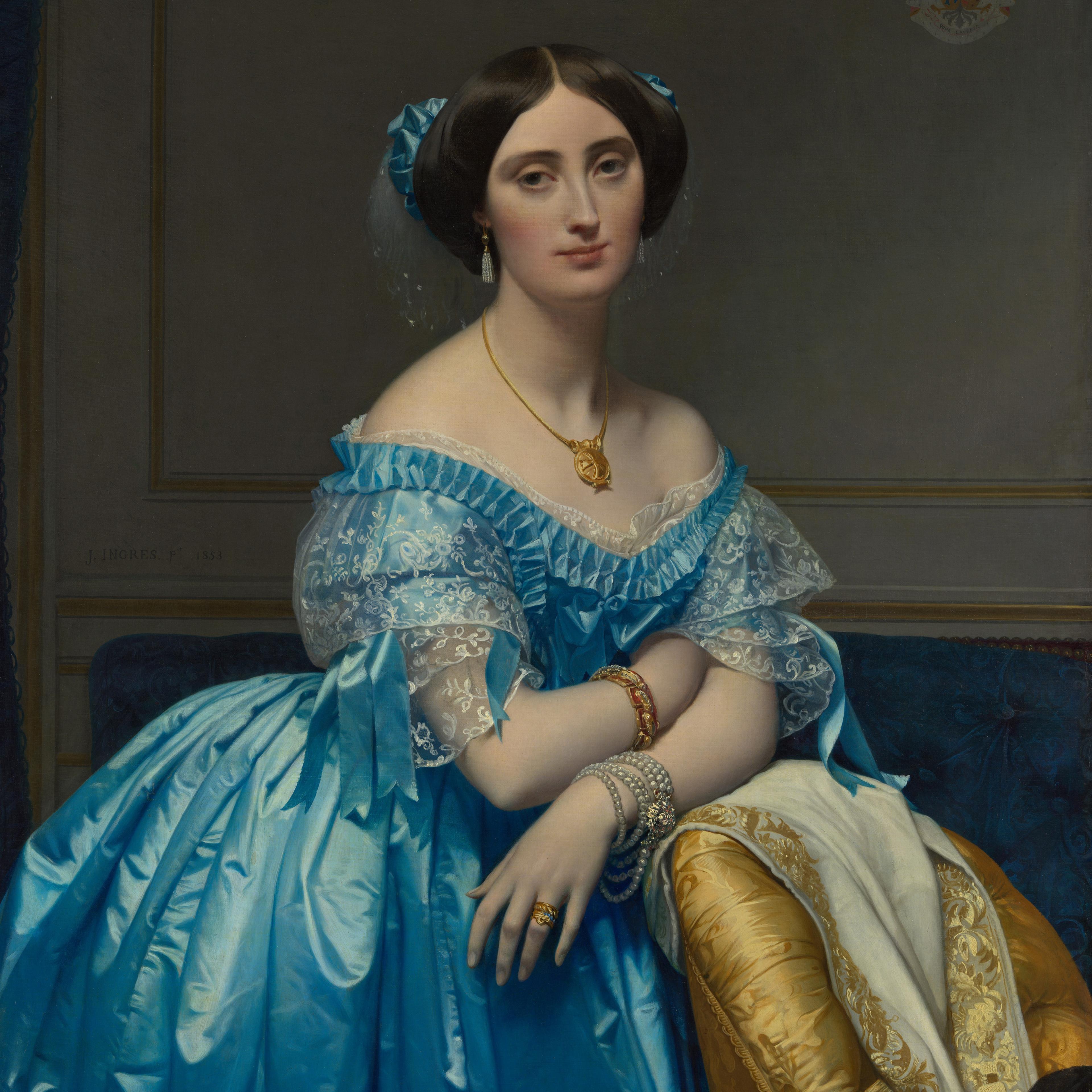 Close-up detail of a woman's hand resting against a yellow chair. She is wearing gold rings and a multi-stranded pearl bracelet.