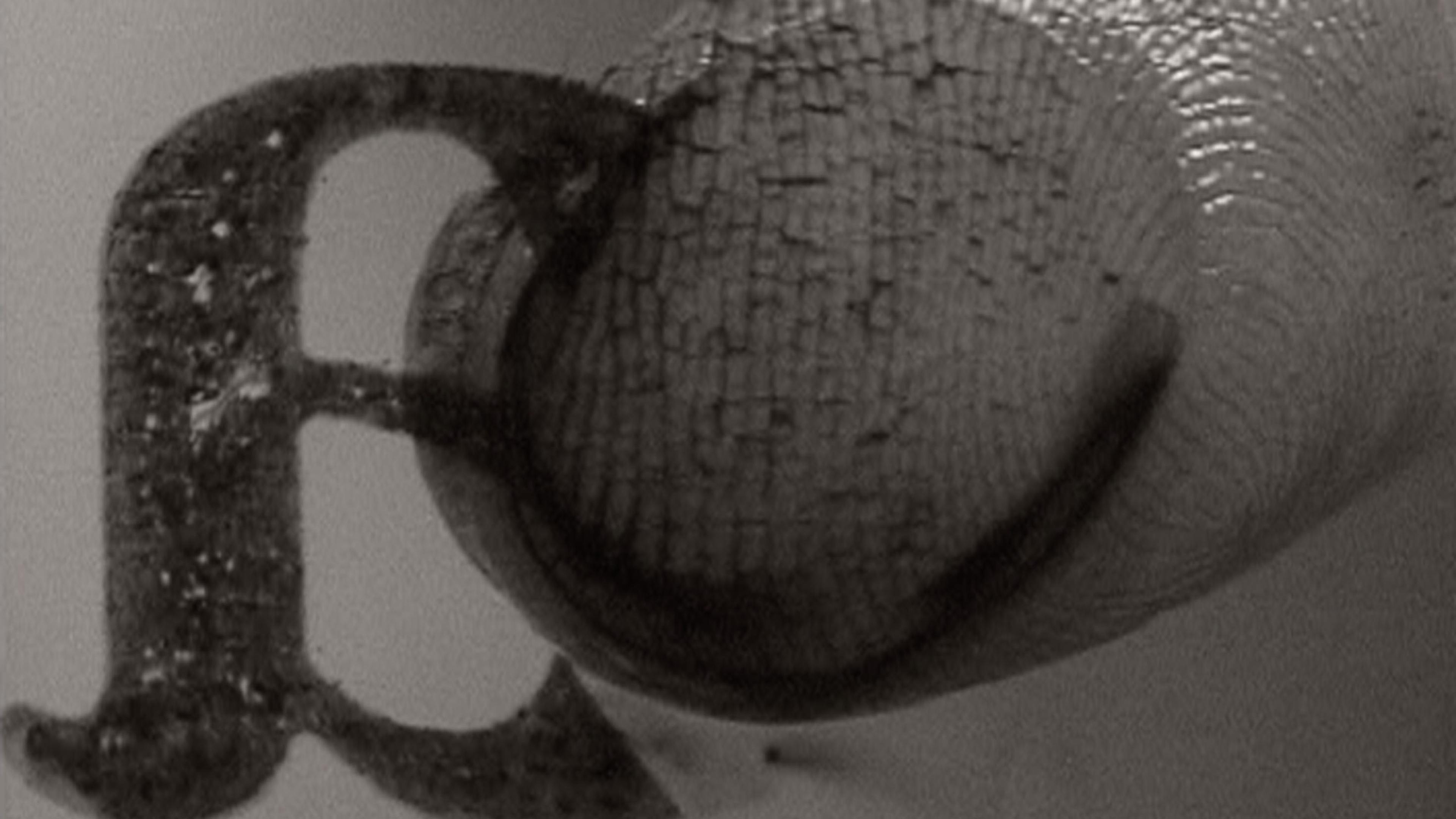 A close-up black and white image of a textured fingertip pressing against a metal printing letter "a" in reverse relief.