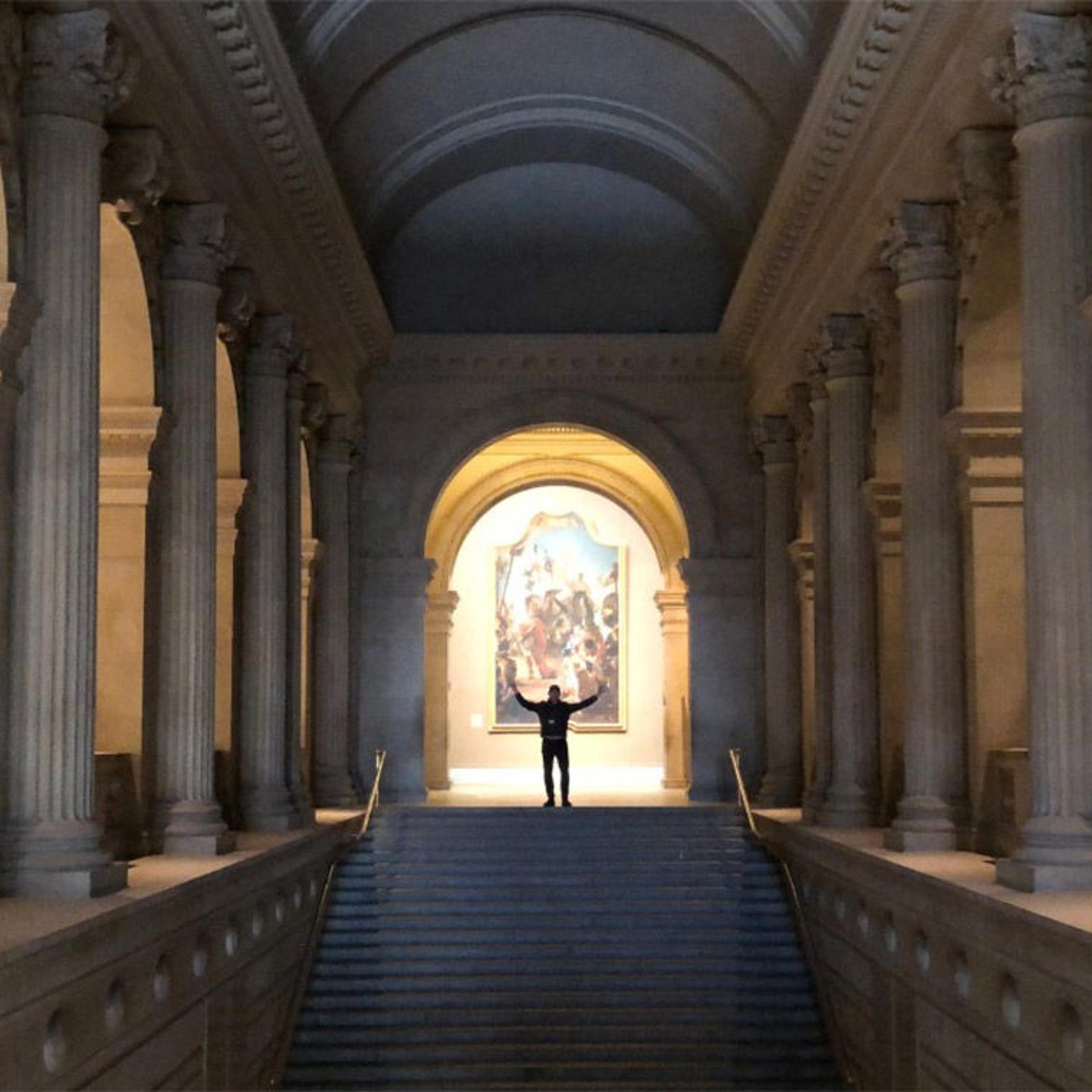 A man stands with his arms outstretched at the top of a large stairwell, surrounded by columns in the Metropolitan Museum of Art