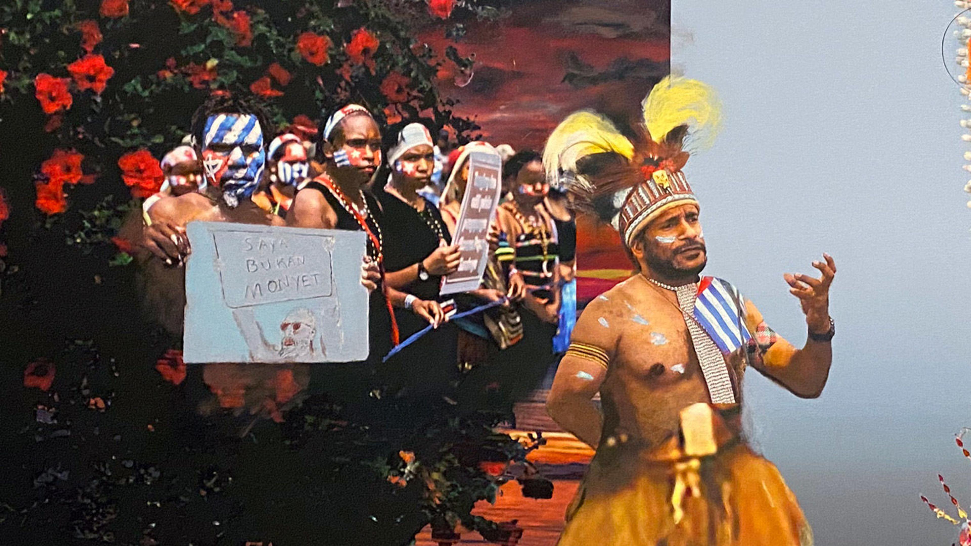 A crowd of people to the left hold up a sign while a shirtless man holds his left hand up as in an oratory gesture