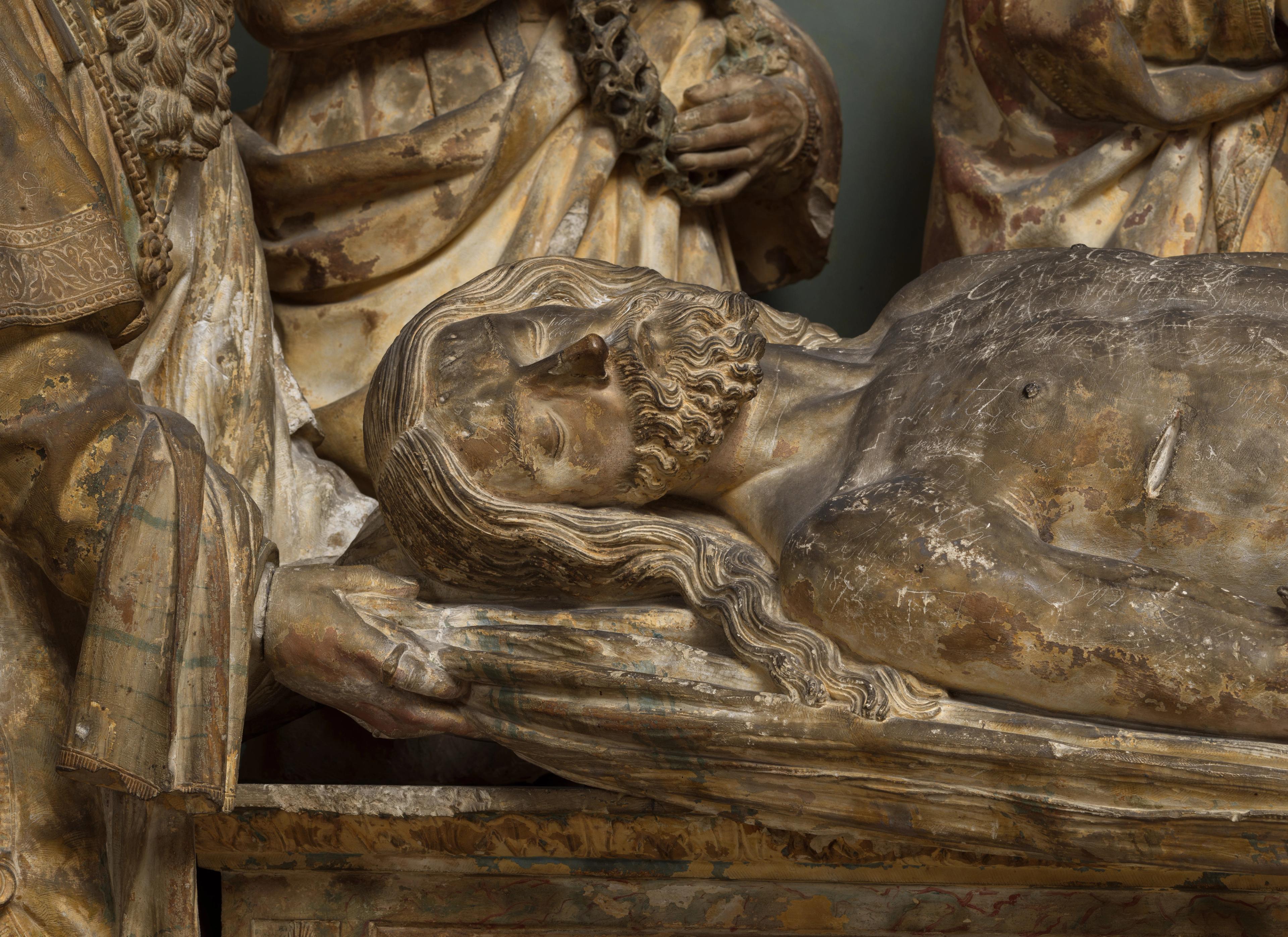 Torso of Christ lying prone on top of a tomb. The close-up of his stone surface reveals varying shades of gray discoloration.
