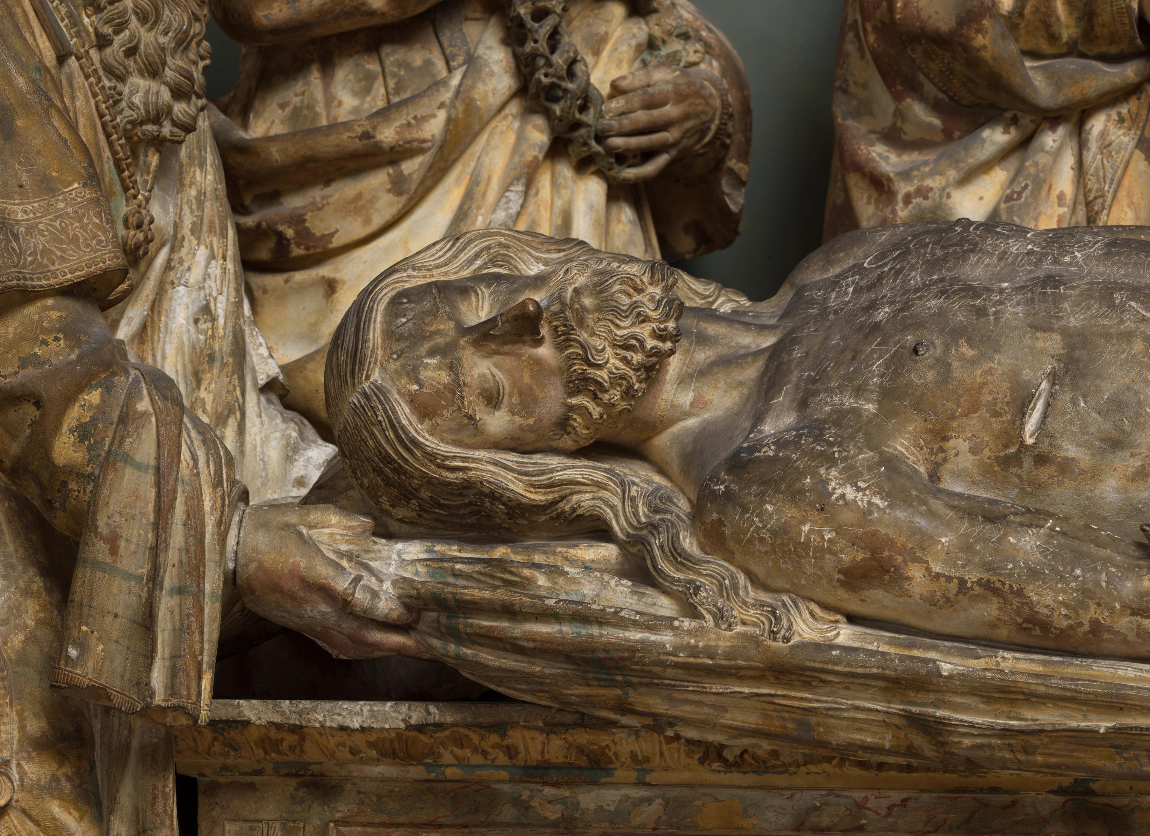 Torso of Christ lying prone on top of a tomb. The close-up of his stone surface reveals varying shades of gray discoloration.