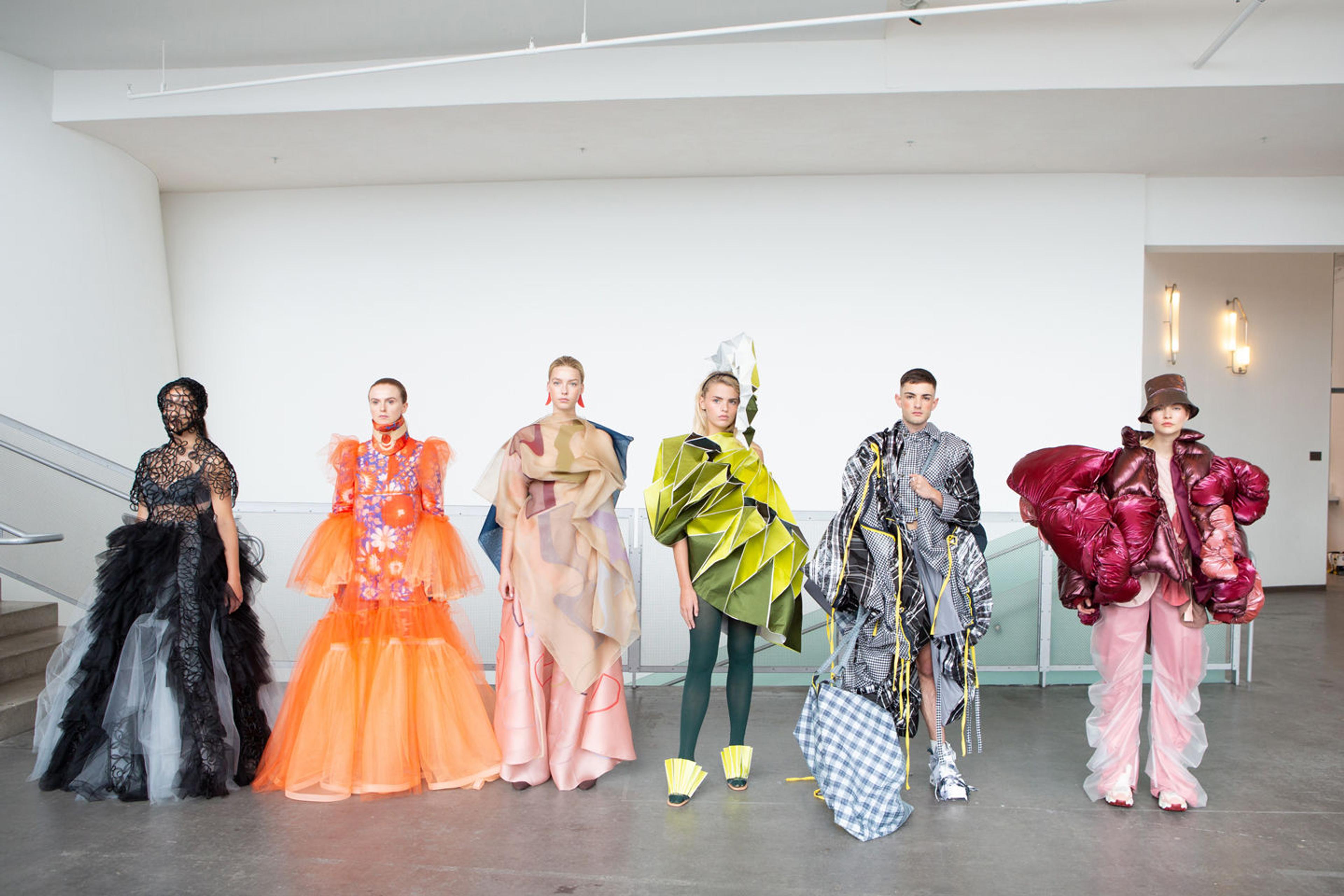 Six models stand in a row within a brightly lit room modeling experimental clothing designs ranging in bright neon colors and complex pleated volumes.