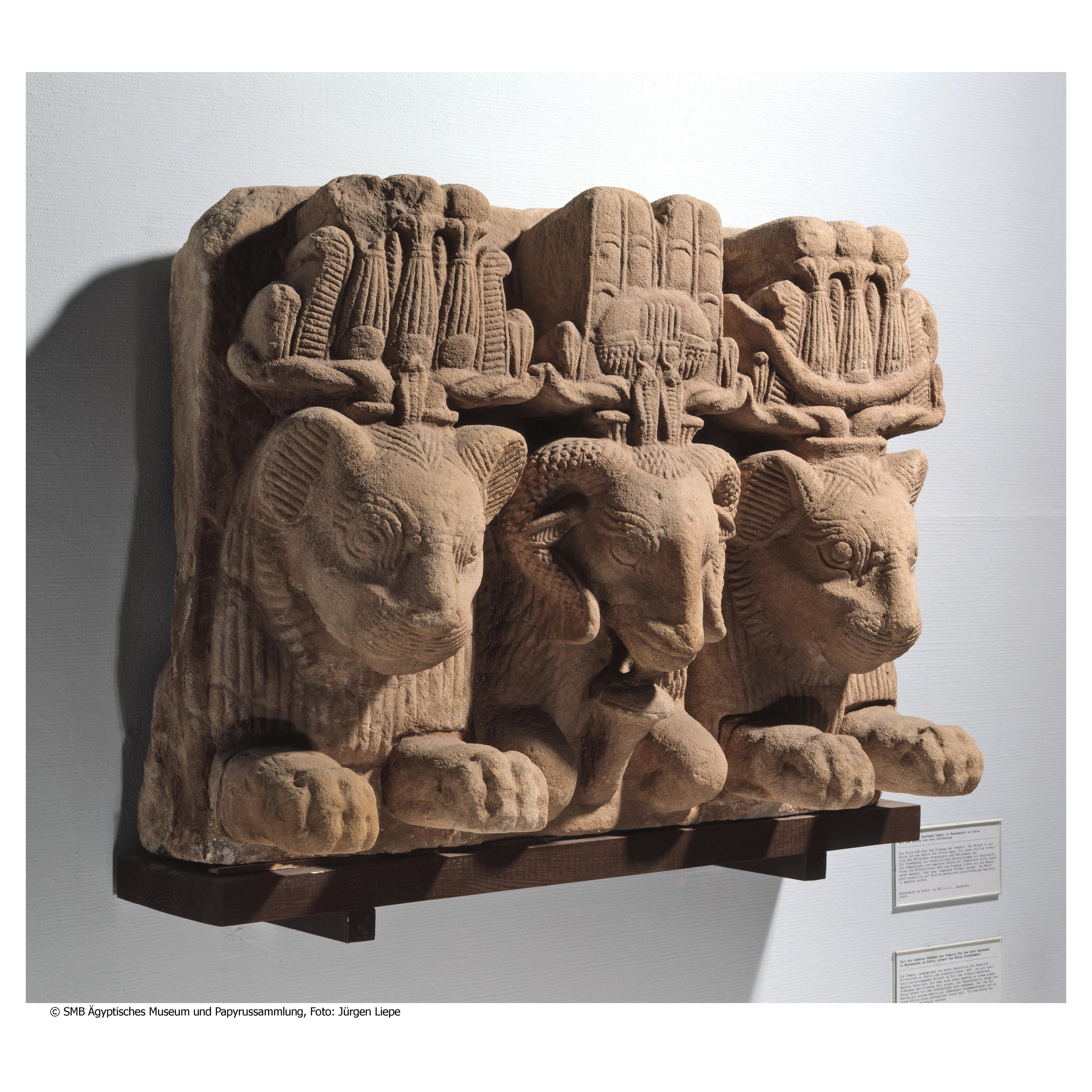 Ancient Egyptian stone relief with two lion heads flanking a ram head, adorned with detailed headdresses.