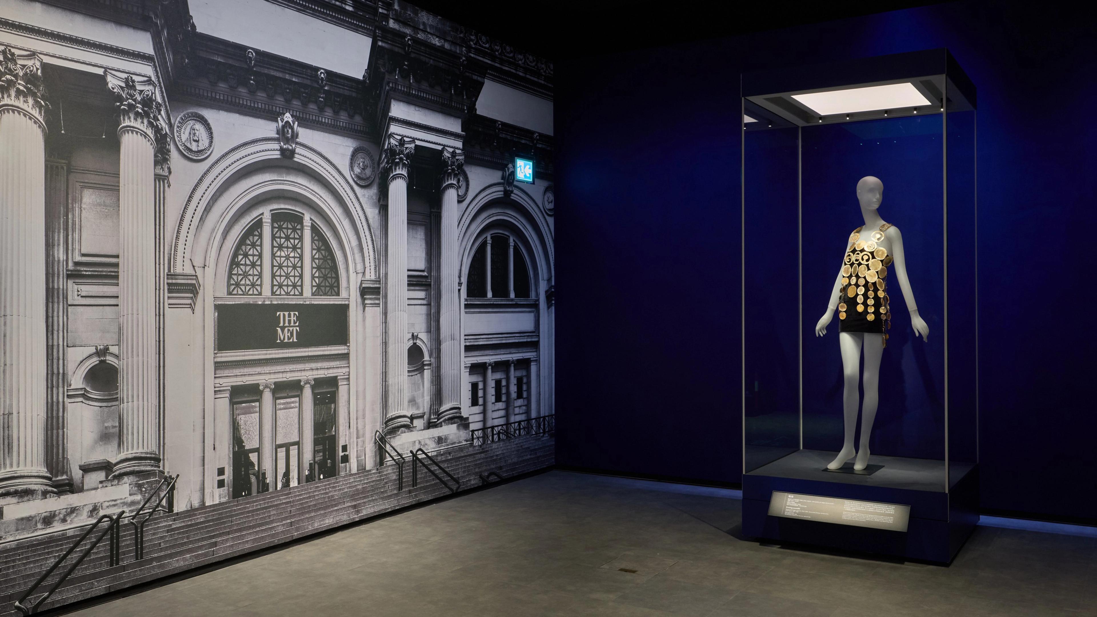 Corner of a gallery with a print of The Met's facade covering the entire lefthand wall, and a vitrine with a mannequin wearing a short black dress adorned with gold coins against the righthand wall.