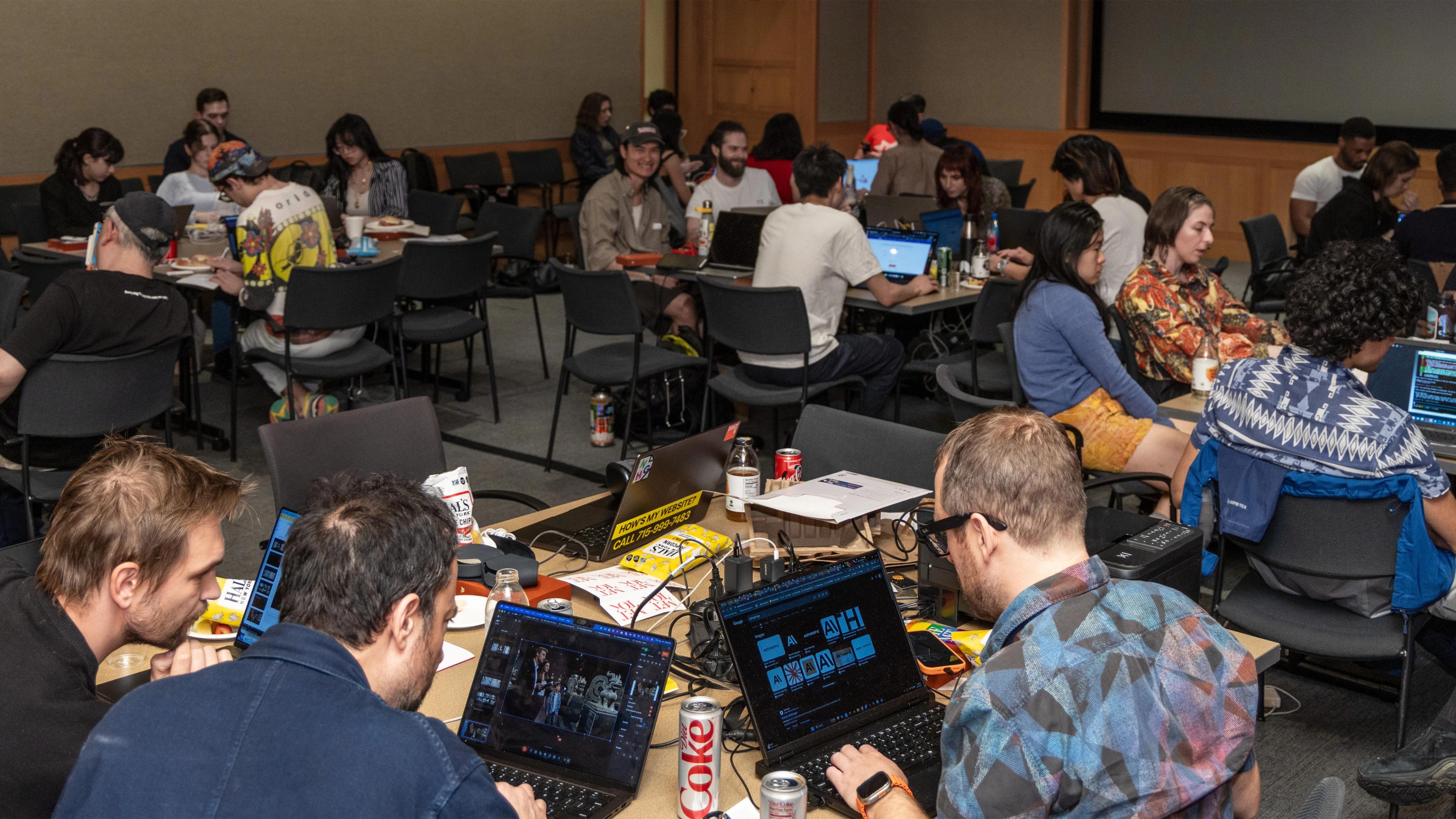 A group of people in a conference room are engaged in collaborative work on laptops. The atmosphere is focused and lively, with drinks and snacks on tables.