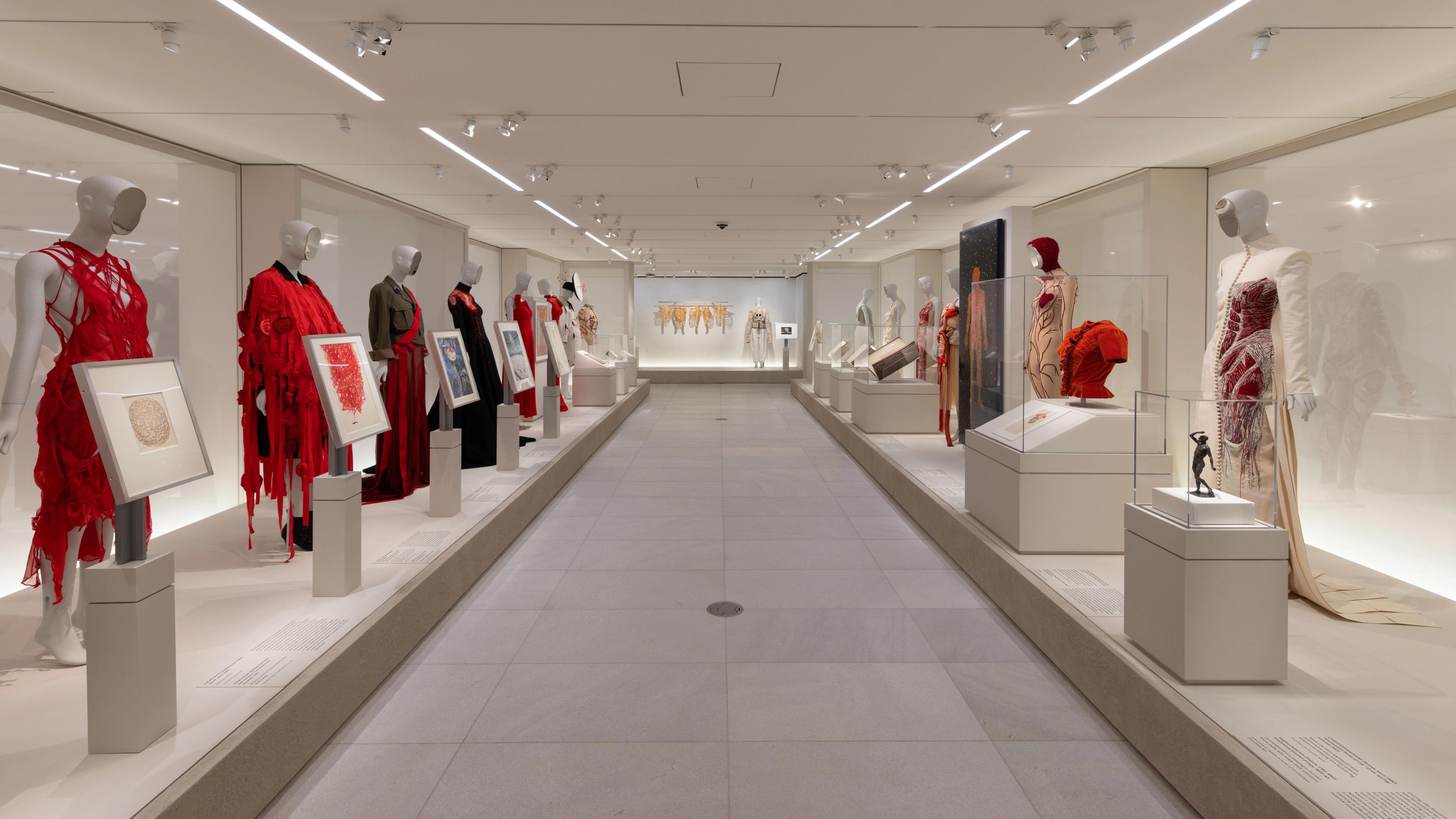 Mannequins in diverse outfits, including a striking red dress, are displayed in a well-lit museum.