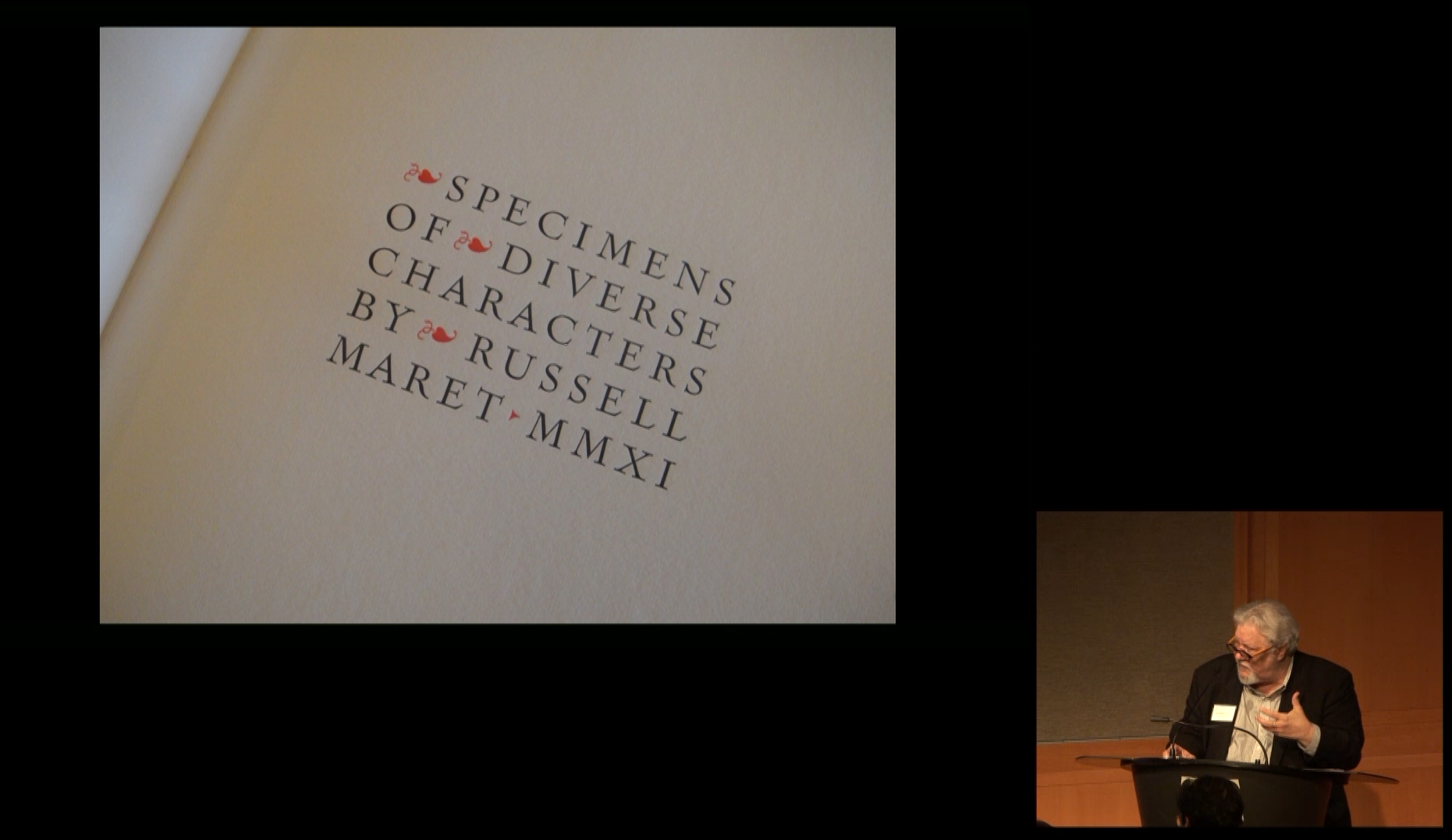 A man speaking at a podium on the right side of a stage with a large projection behind him showing a white piece of paper with the title and author of a book printed in black letters. 