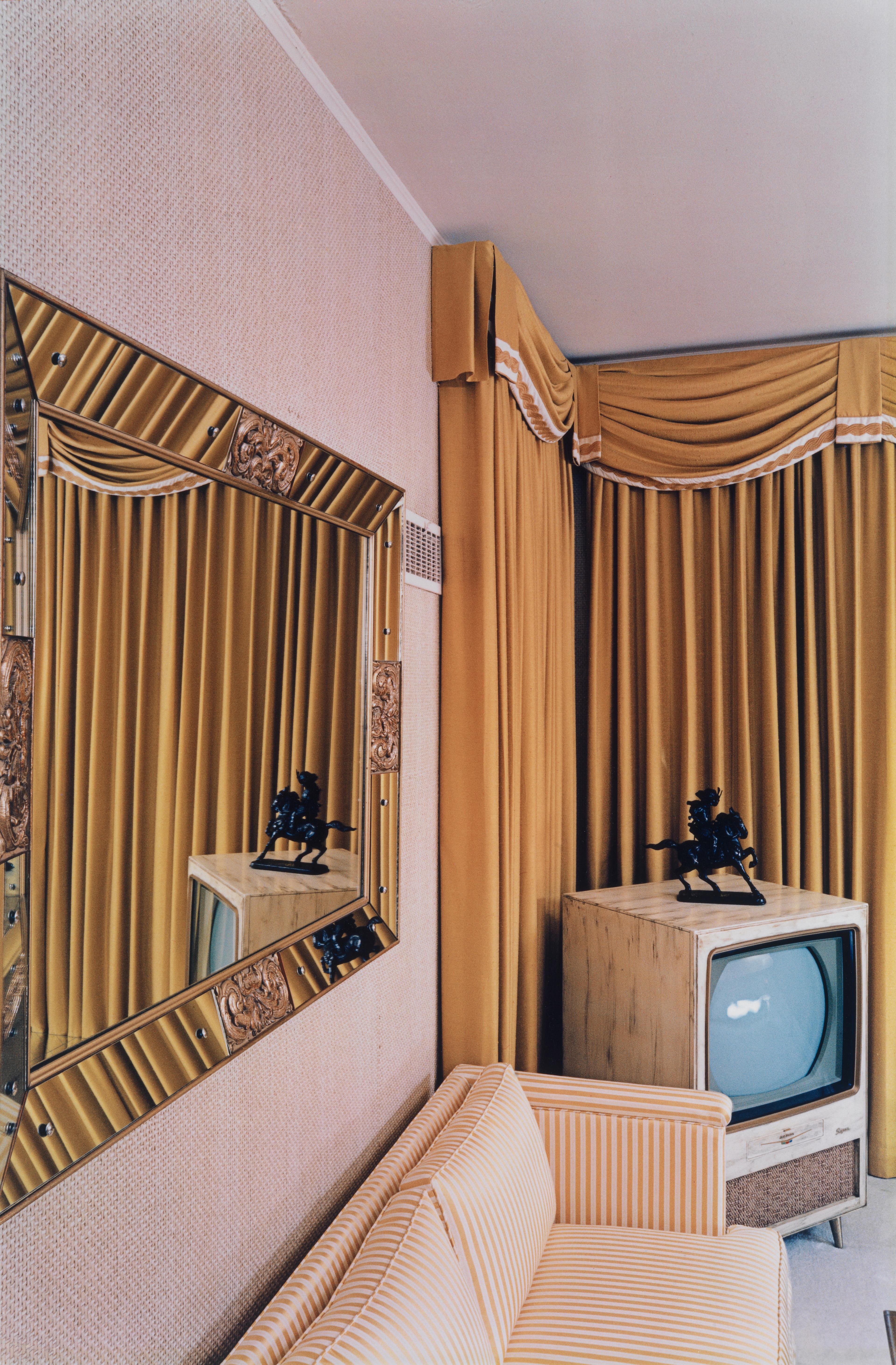 A color photograph of a room with a striped sofa, an ornate mirror reflecting elaborate drapery, and a TV set.