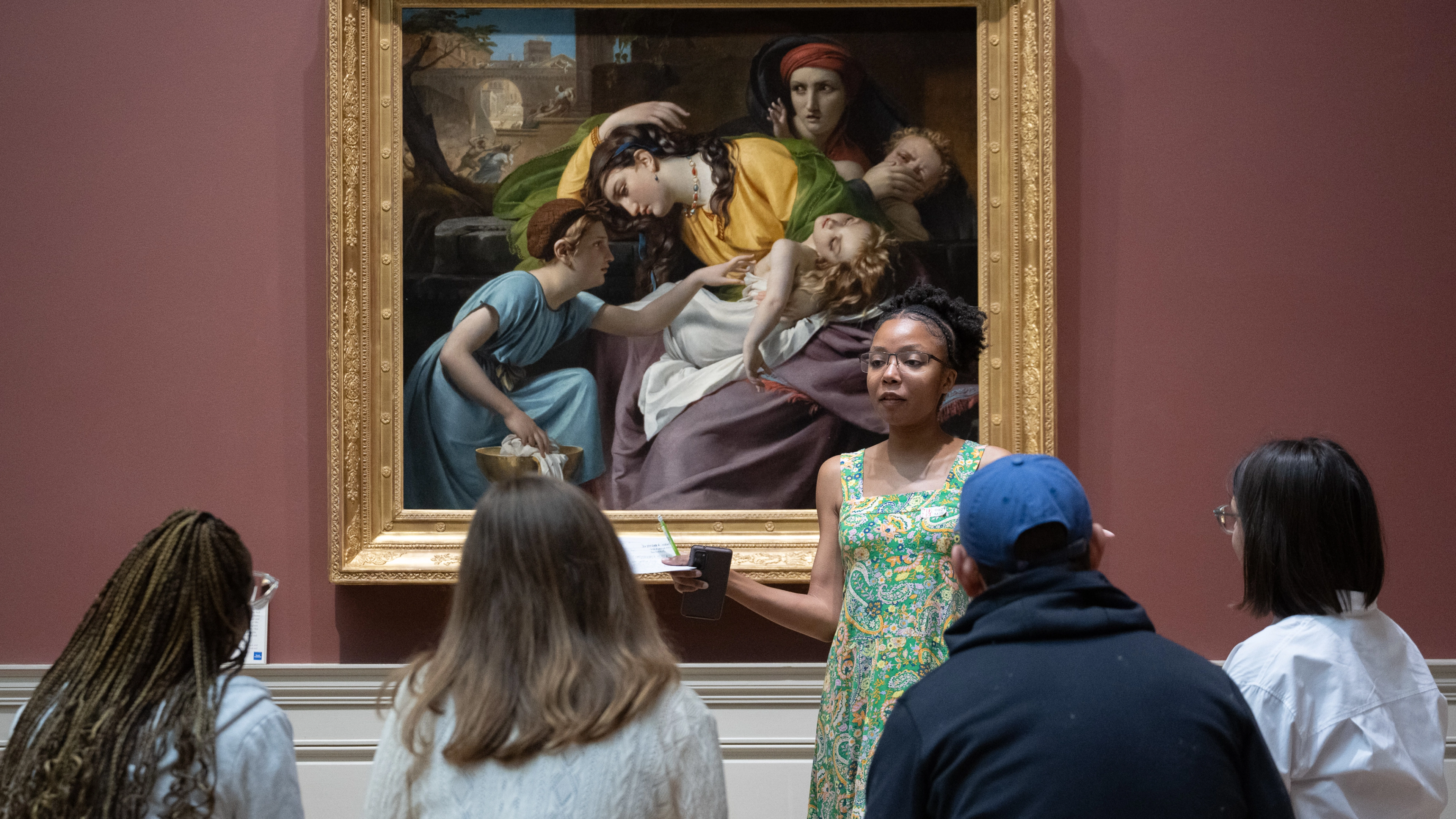 A group of people sit on a bench in front of an artwork. The person in the middle is discussing the artwork.