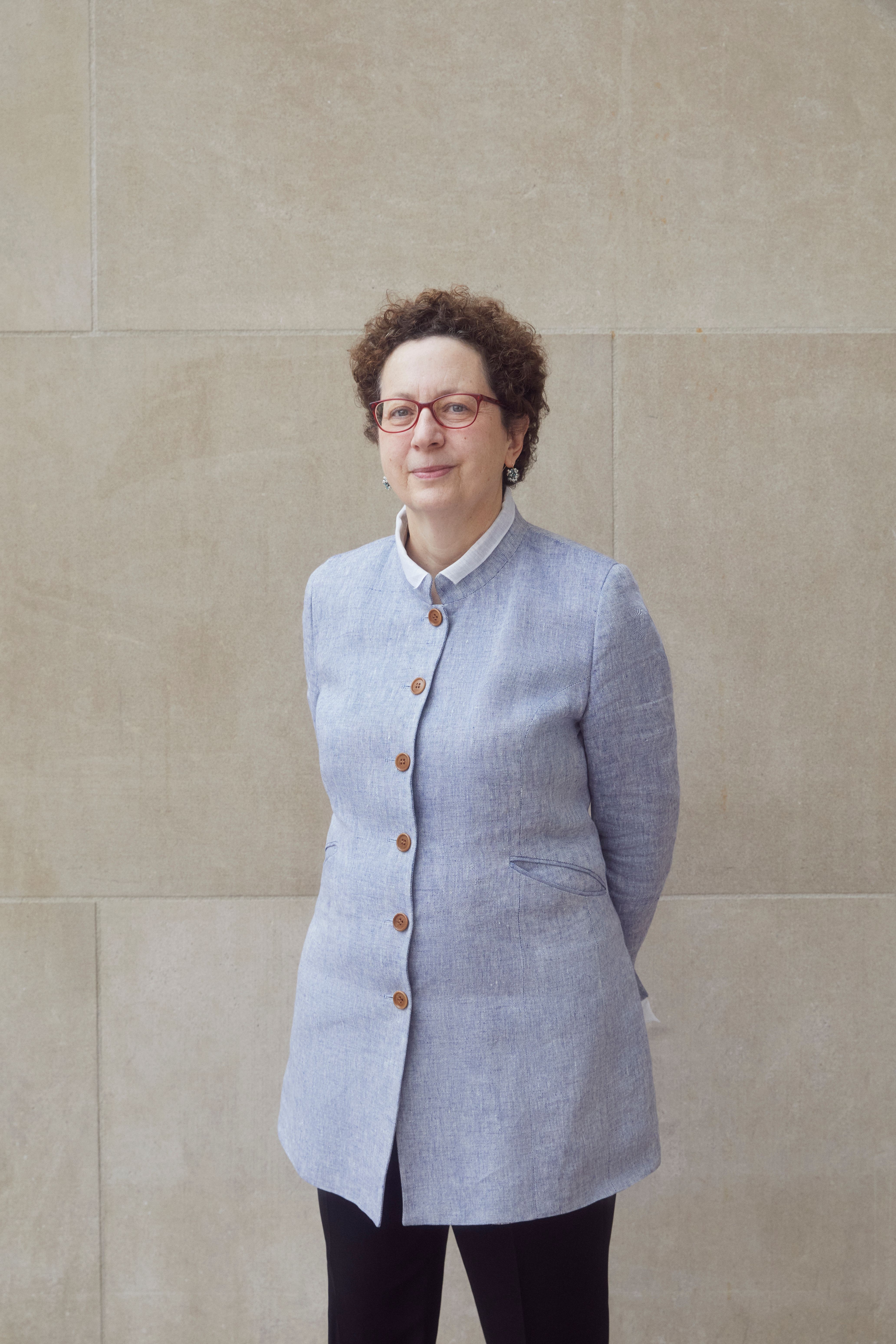 A color photograph of Andrea Bayer standing in front of a stone wall in a long light blue jacket.