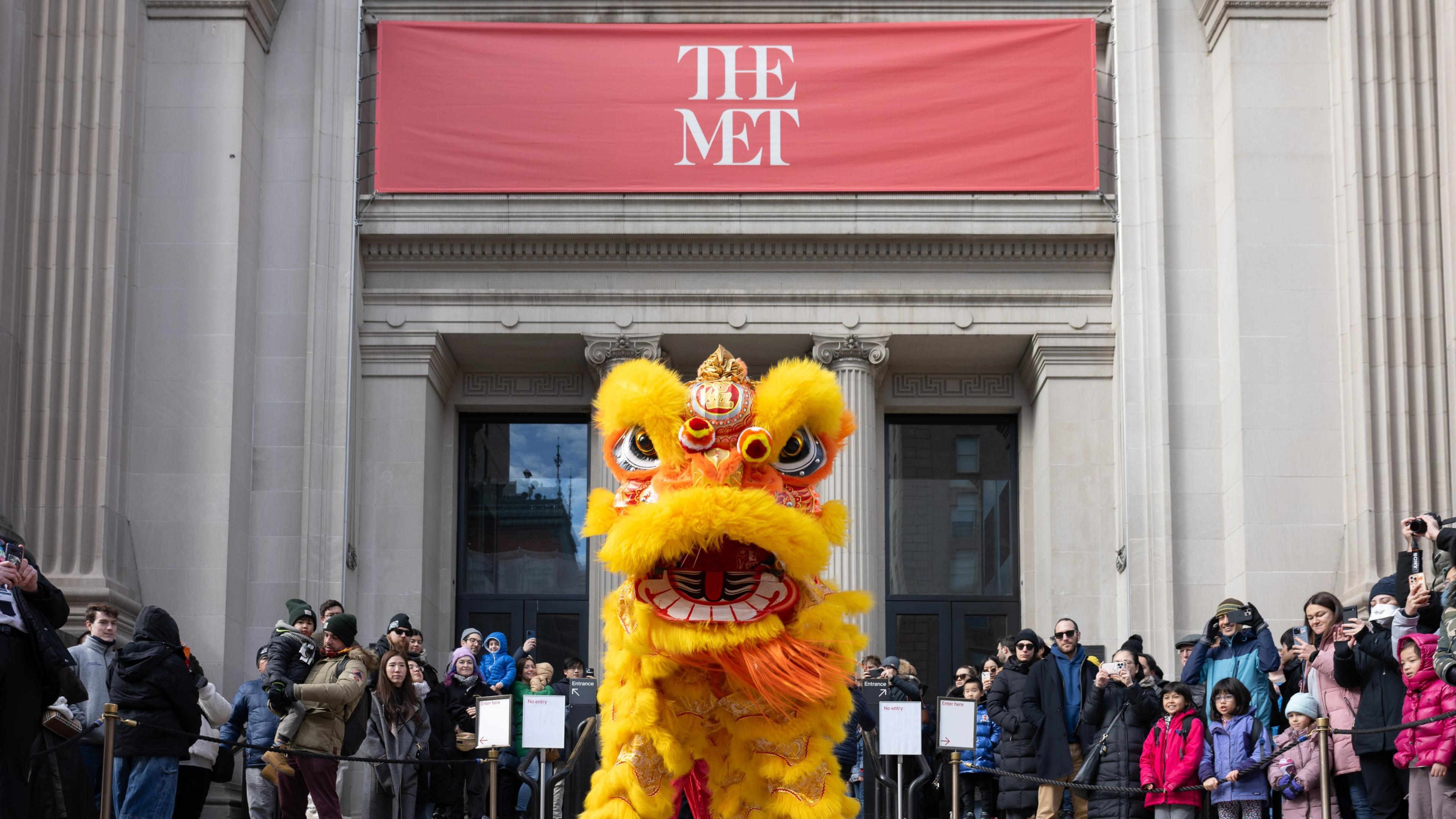 A lion costume in front of a building with a banner atop saying "The Met"