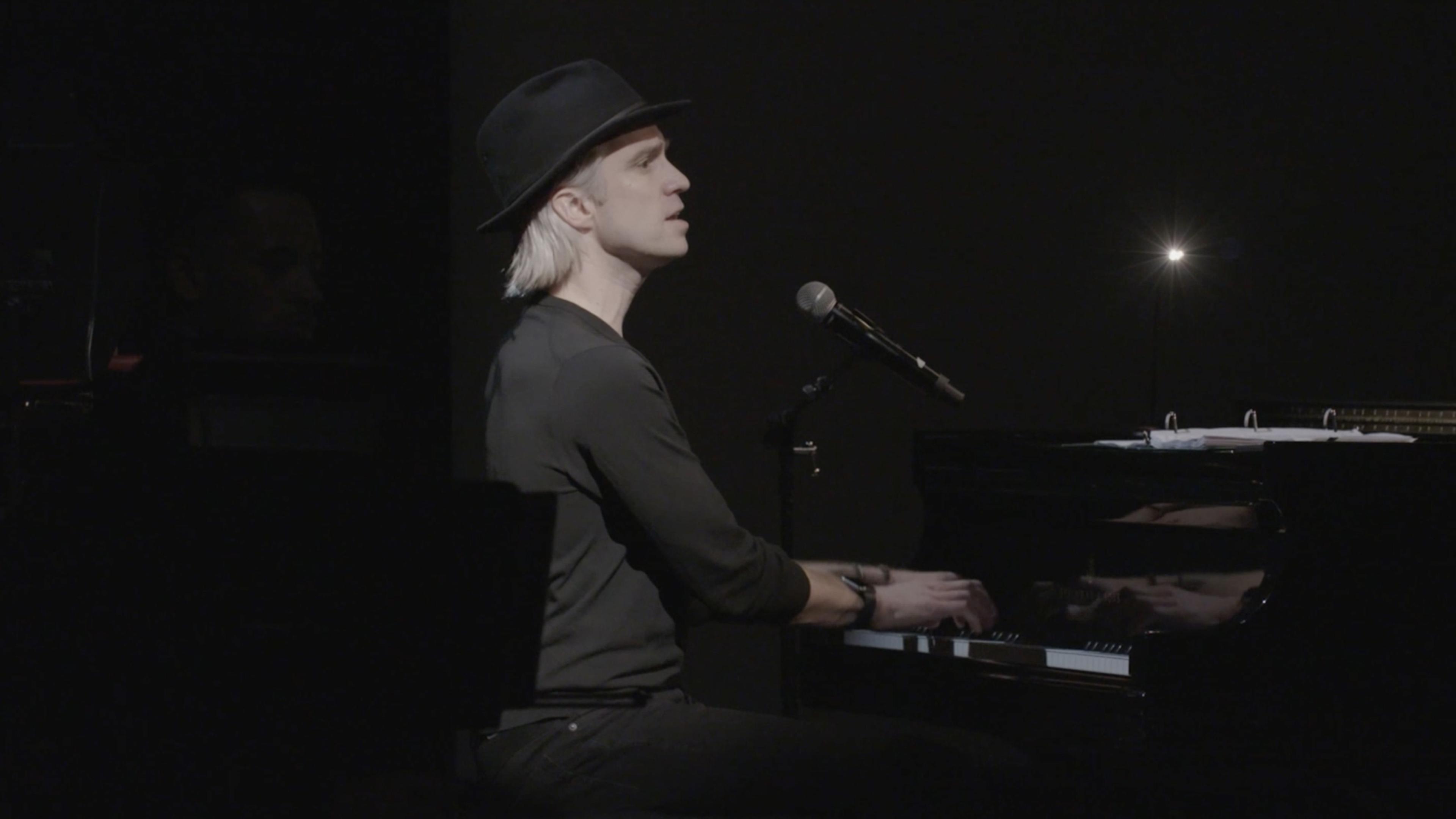 Gavin Creel singing at the piano, wearing a black hat and shirt against a black background.