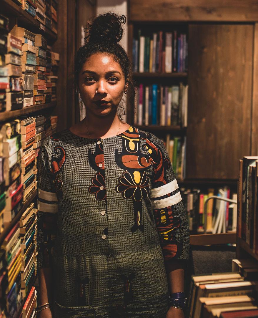 Image of a Black women in front of a small library of books on wooden shelves. The women's black hair is up in a bun and the yellow toned lighting creates a shadowy effect. She is wearing a patterned outfit with buttons and has dark lipstick with a nose ring. She is staring directly into the camera from the waist up.