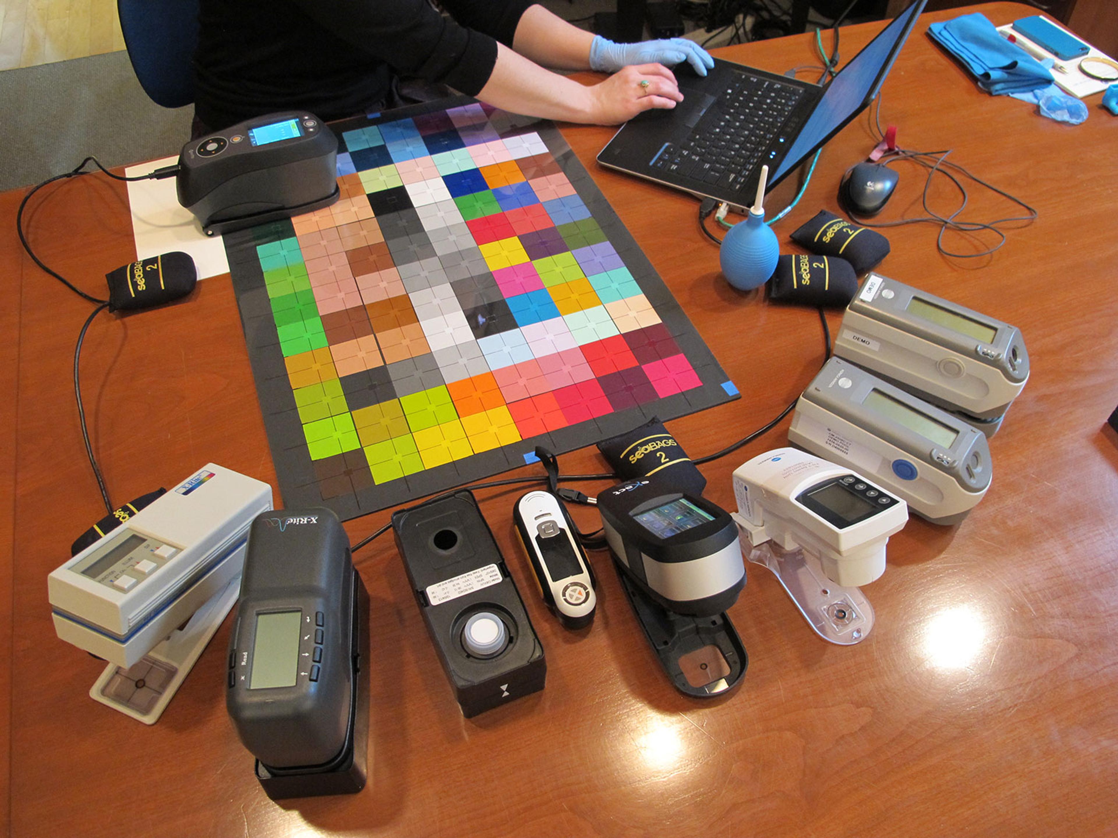 An overhead view of a someone typing on a laptop at a large wooden table surrounded by a multi-color grid mat and various electronic devices.