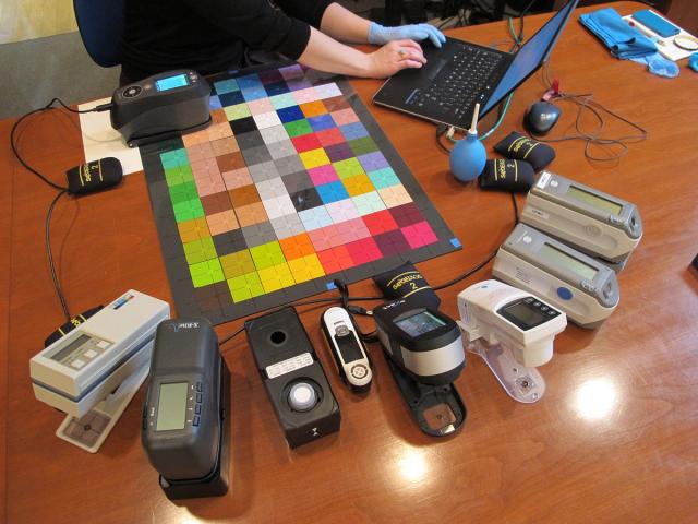 An overhead view of a someone typing on a laptop at a large wooden table surrounded by a multi-color grid mat and various electronic devices.