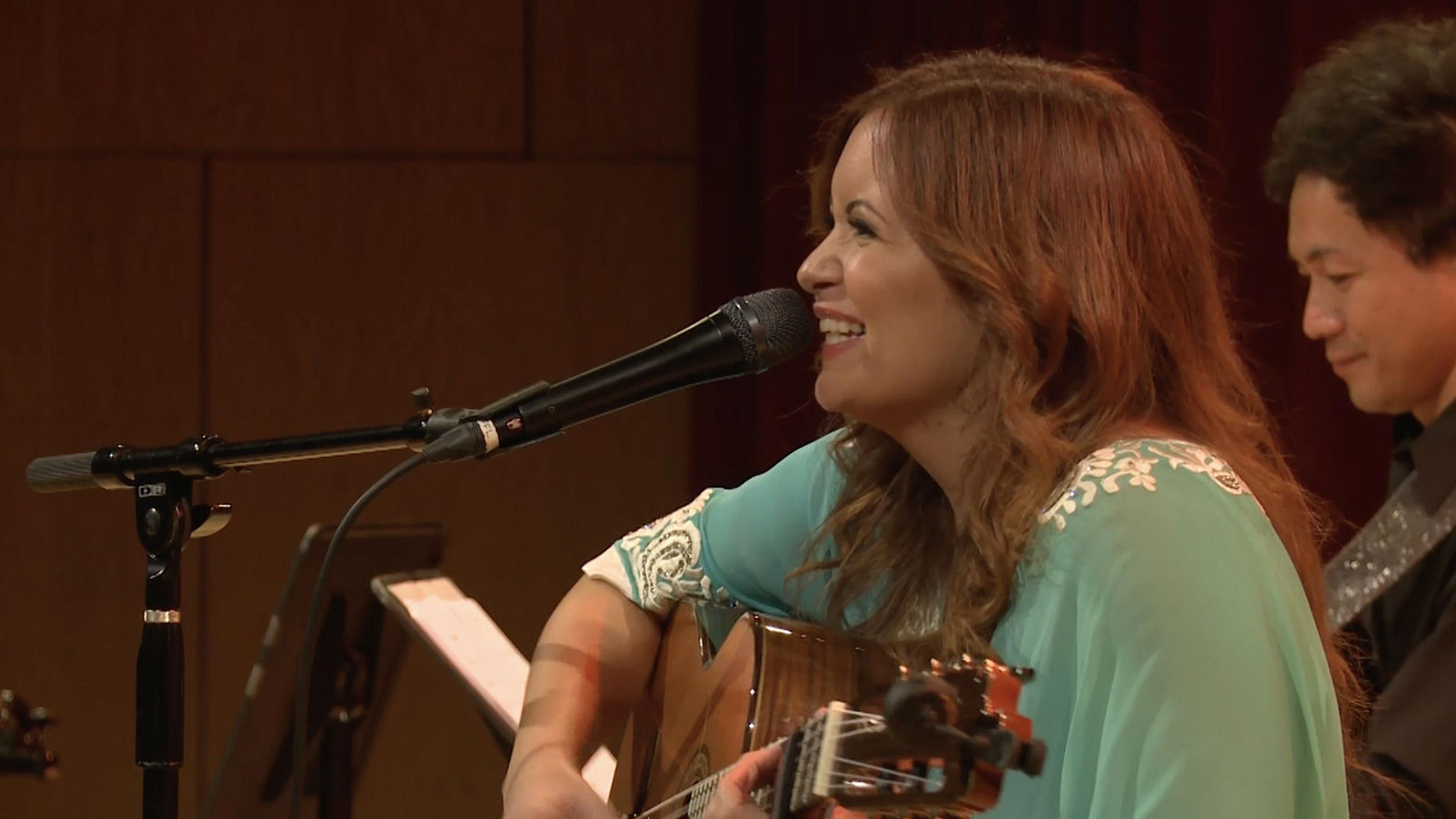 Profile view of Farah Siraj smiling, while sitting with a guitar in front of a microphone.