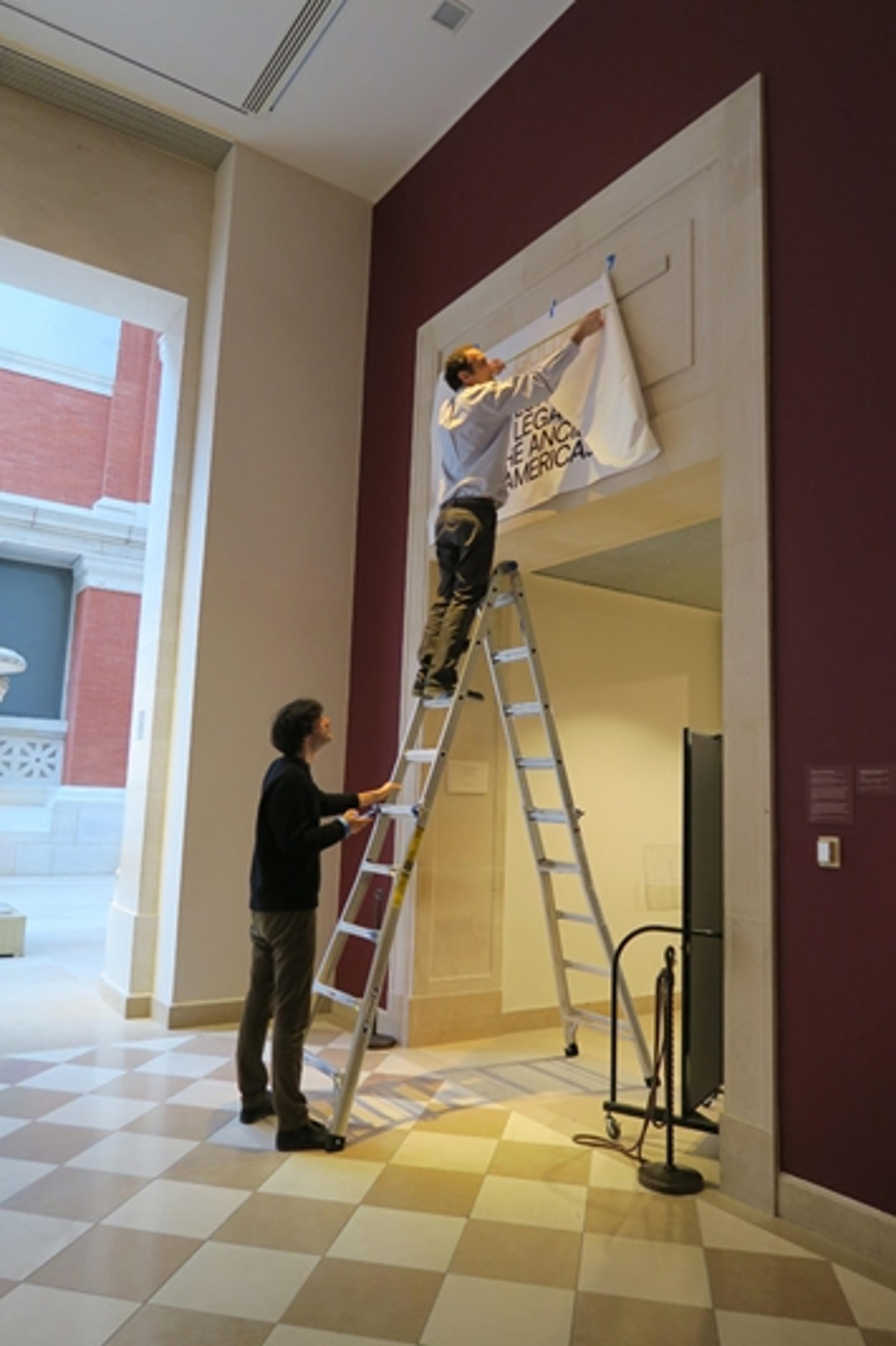 Two Met staff members tape a mock-up of an exhibition design to a gallery wall