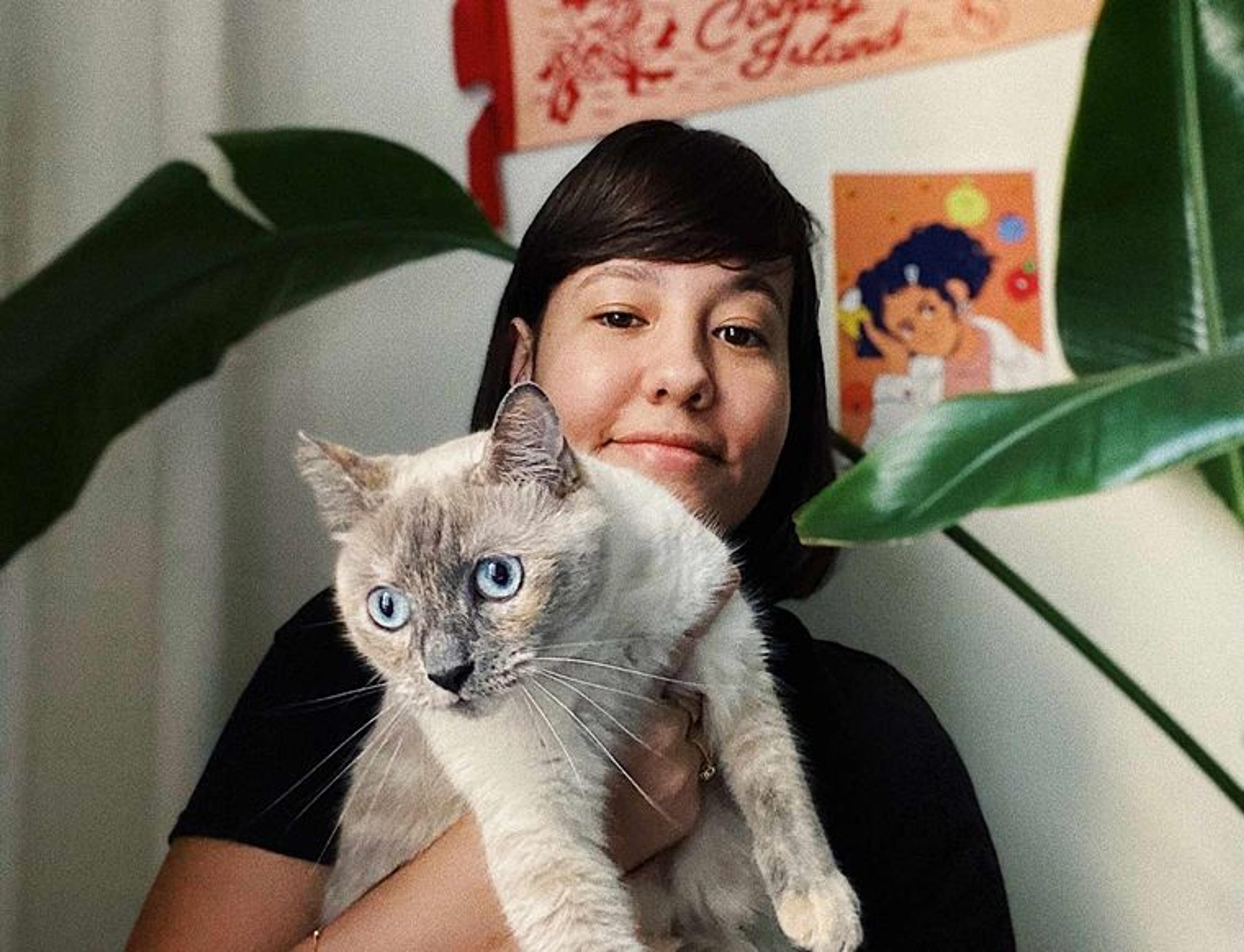 A smiling, dark-haired woman holds up a gray cat while facing the camera in a room surrounded by posters and the leaves of houseplants.