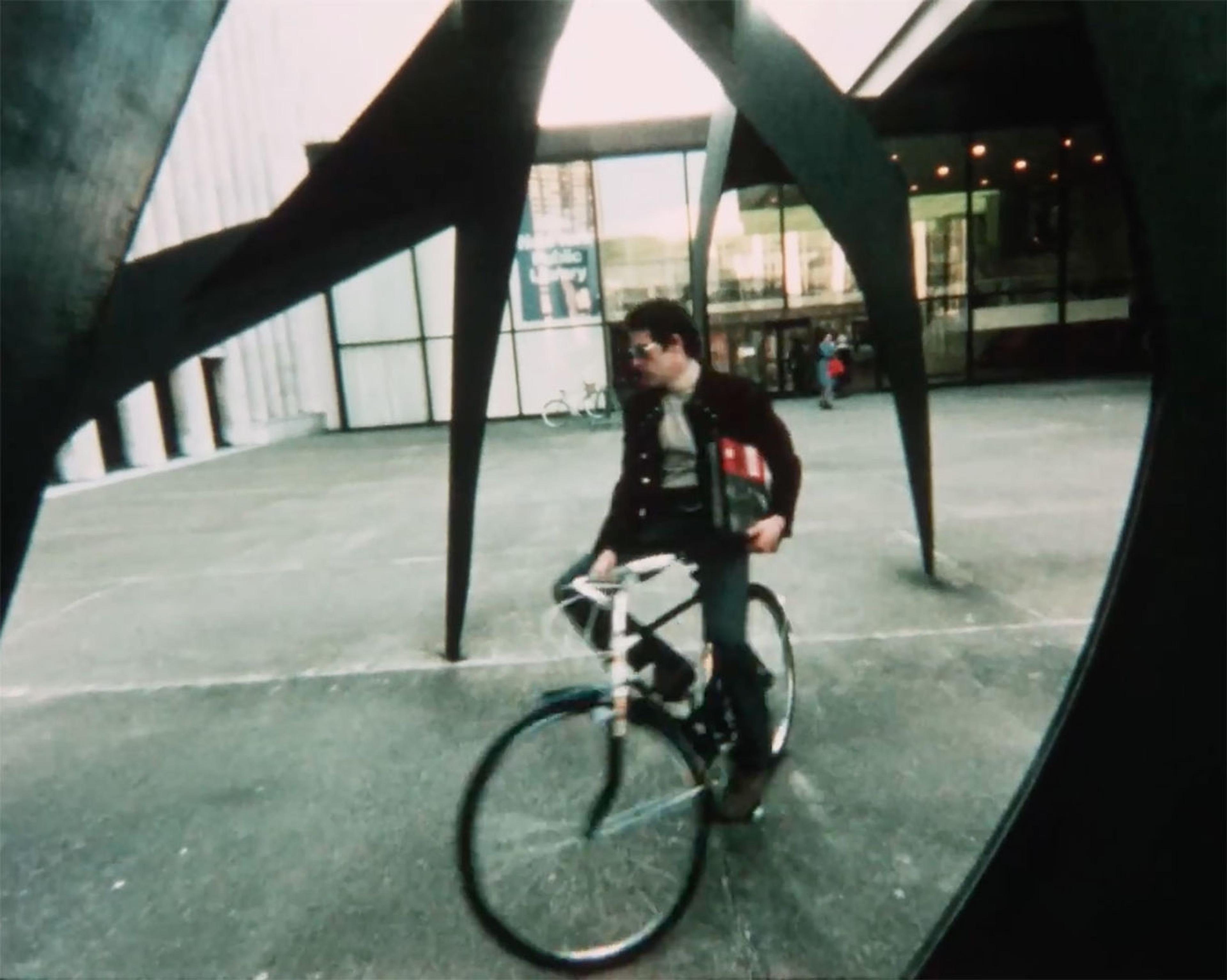 A photo of a man riding his bike in a city underneath a steel sculpture