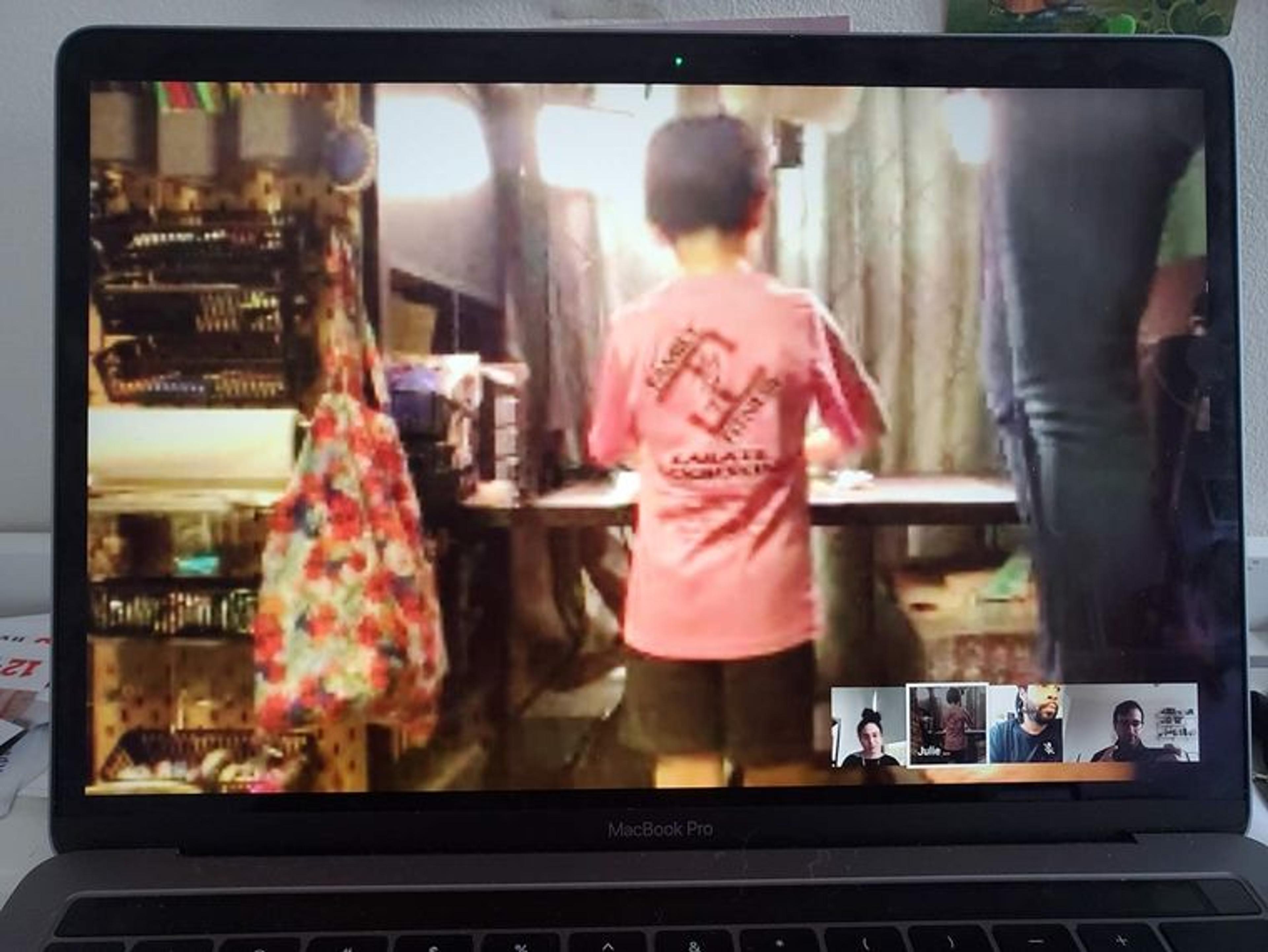 A laptop displays a video chat of a child in a pink shirt with their back turned, doing something not visible at a table. In the bottom right corner of the laptop screen are chat windows displaying a woman and two men in separate displays.