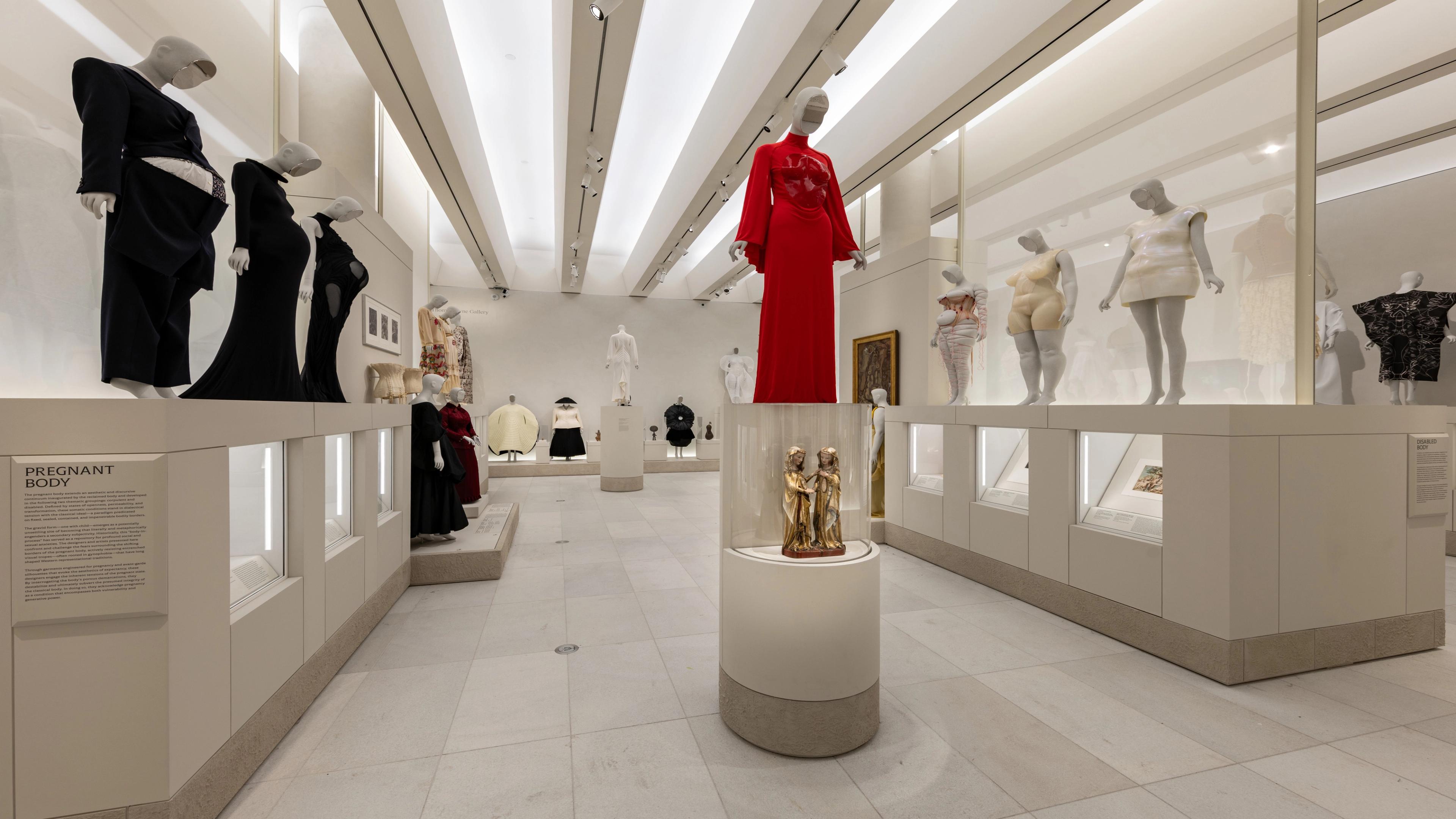 Mannequins in diverse outfits, including a striking red dress, are displayed in a well-lit museum.