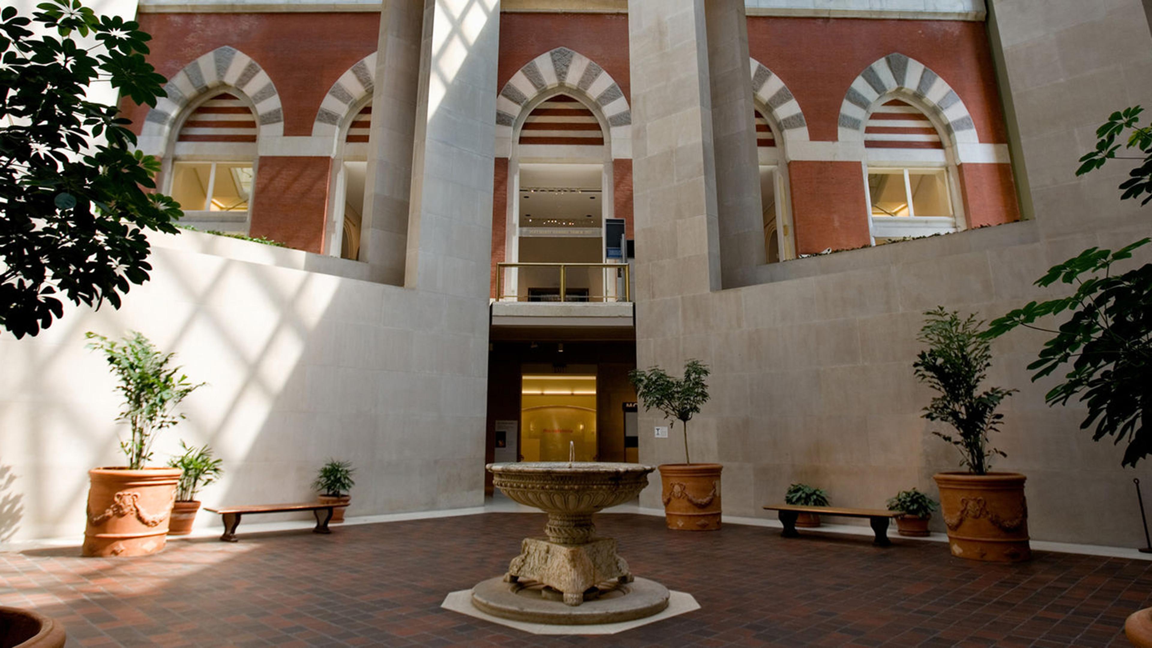 An indoor atrium featuring a stone fountain surrounded by potted plants and benches, with large, bright windows overhead.