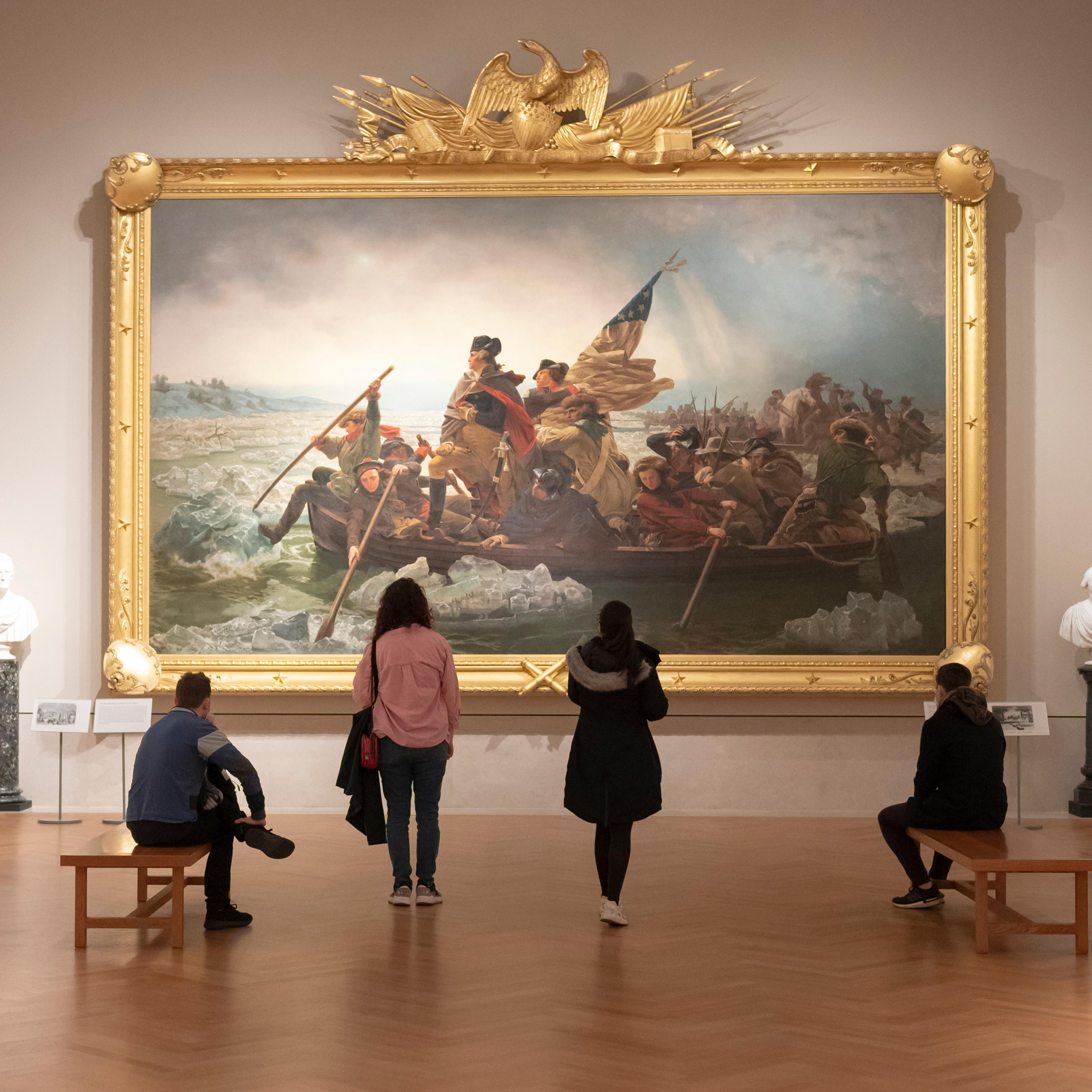People stand in front of a painting where George Washington stands at the bow of a boat as soldiers row across an icy Delaware River under a cloudy sky during the Revolutionary War.