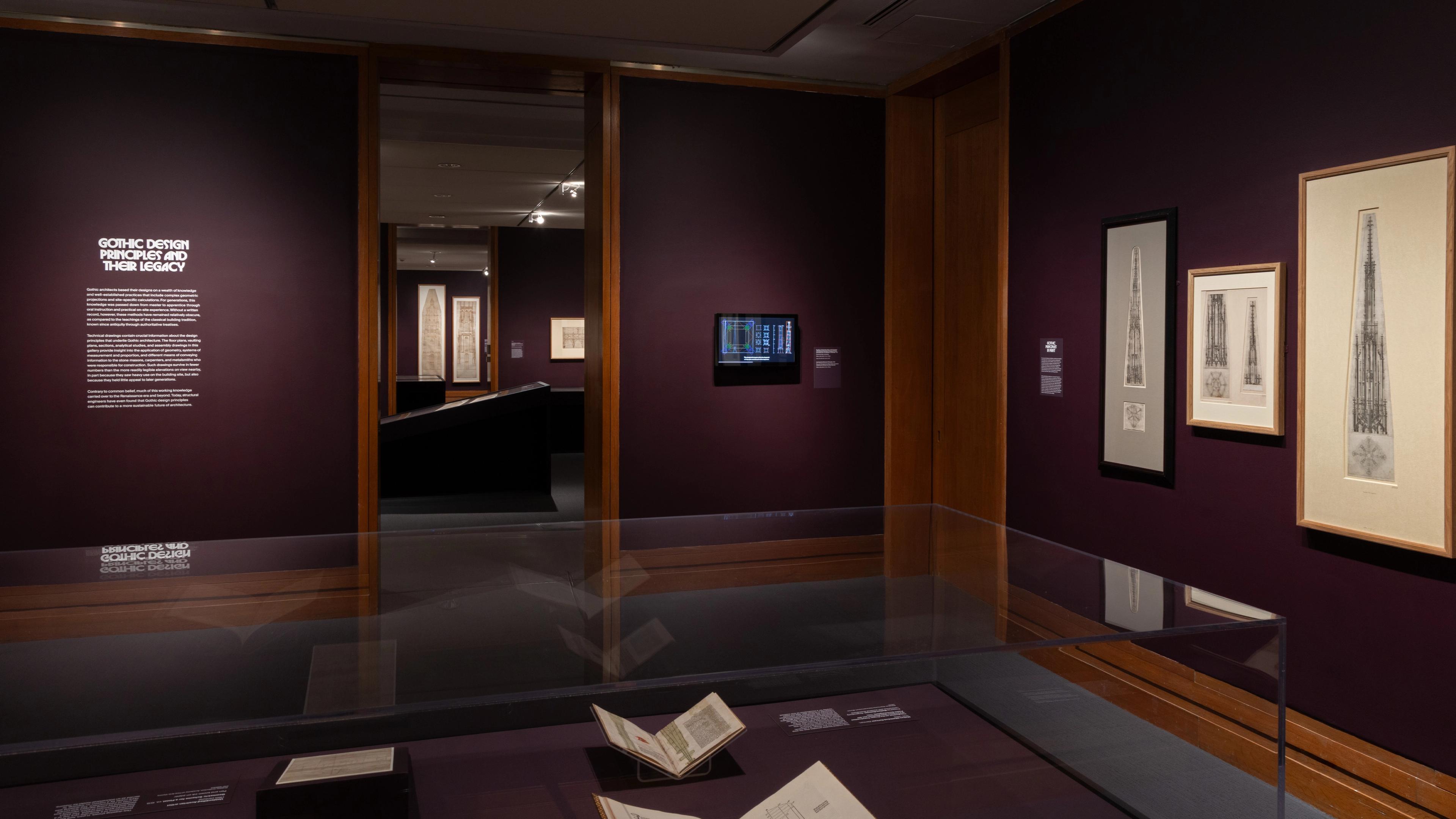 Dimly lit museum exhibit featuring Gothic design artworks on dark purple walls.